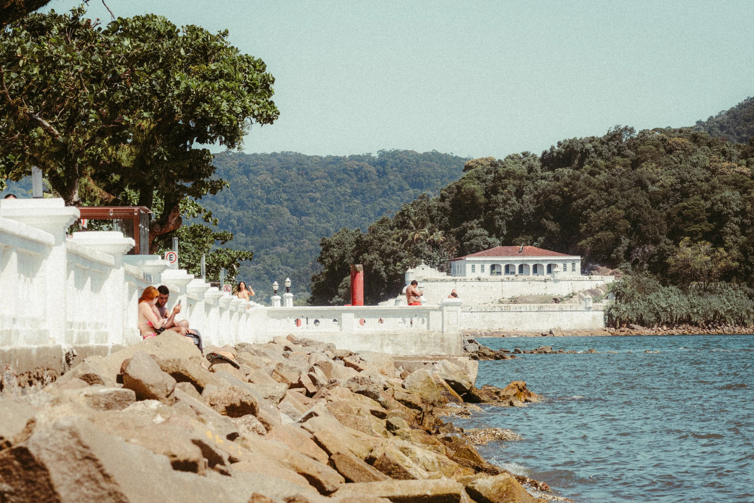 Praia com pessoas sentadas na parede branca ao longo do litoral, árvores, rochas na água, edifícios históricos ao fundo e colinas verdes.