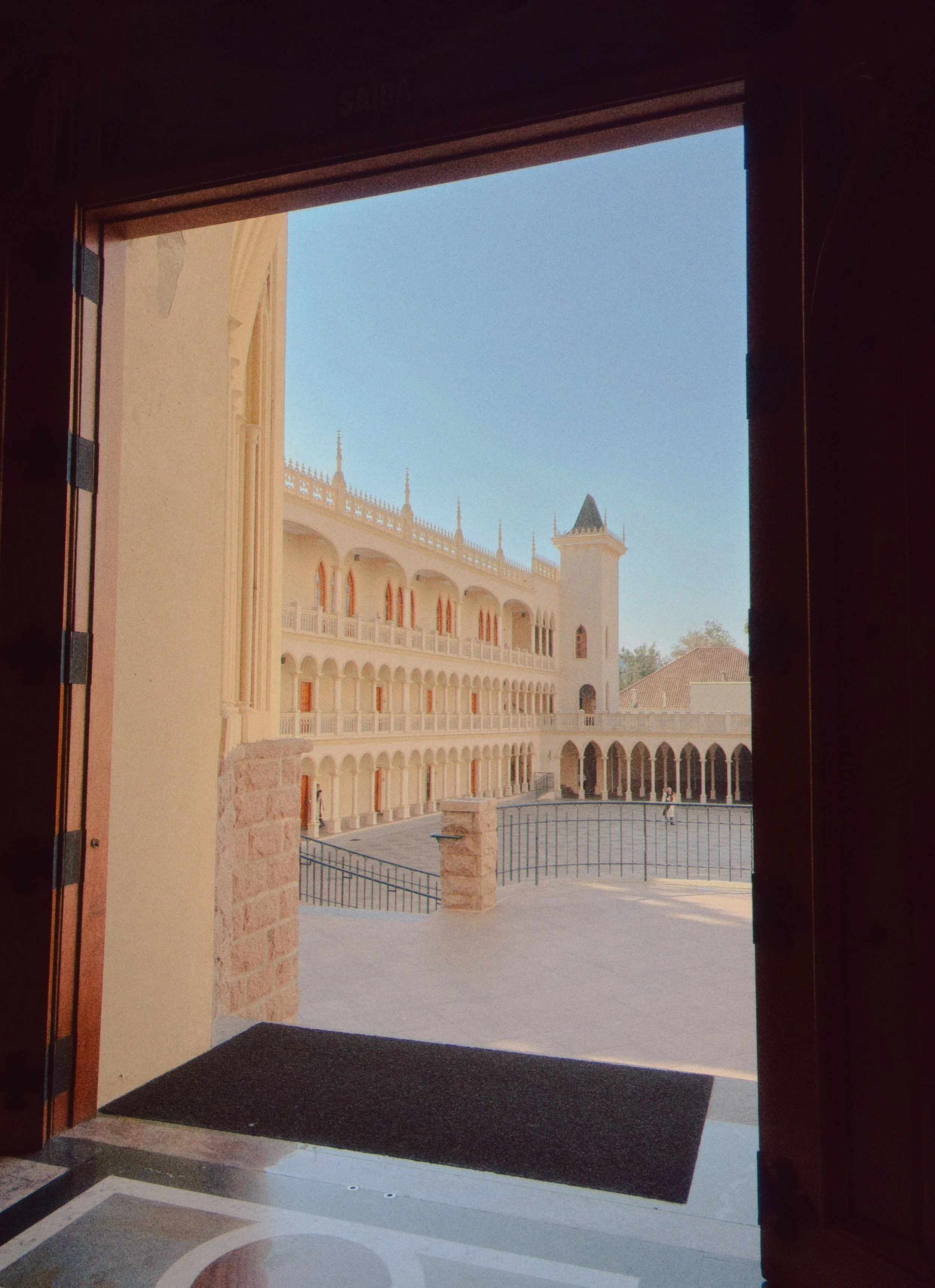 Vista de um pátio interno de um castelo com arquitetura gótica, com vários arcos, torre com espigão, e uma pessoa caminhando ao fundo, vista através de uma porta aberta.