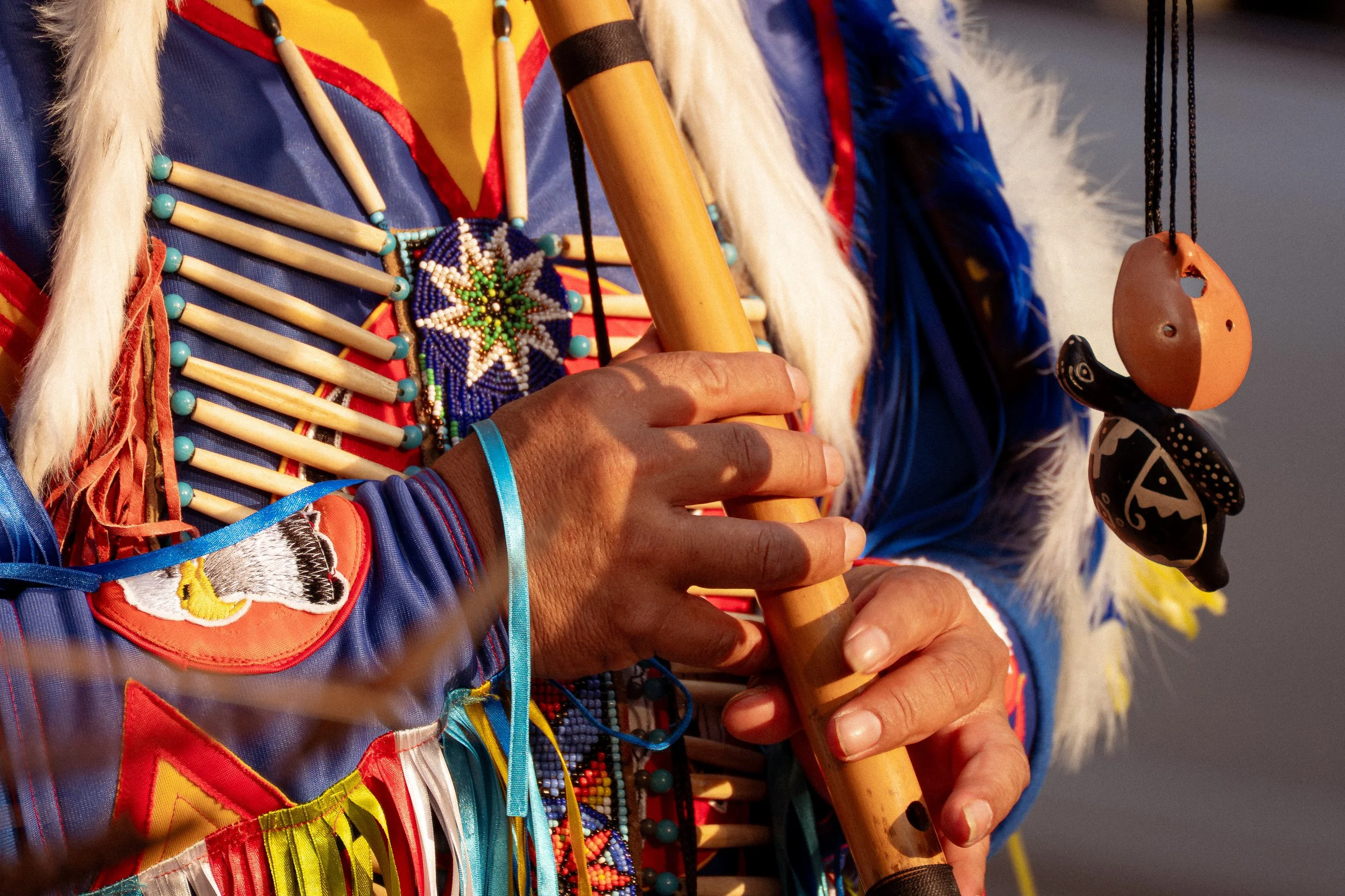 Pessoa indígena vestida com roupas tradicionais coloridas, segurando uma flauta e usando acessórios de miçangas e penas, próximo a um colar de conchas flutuantes.