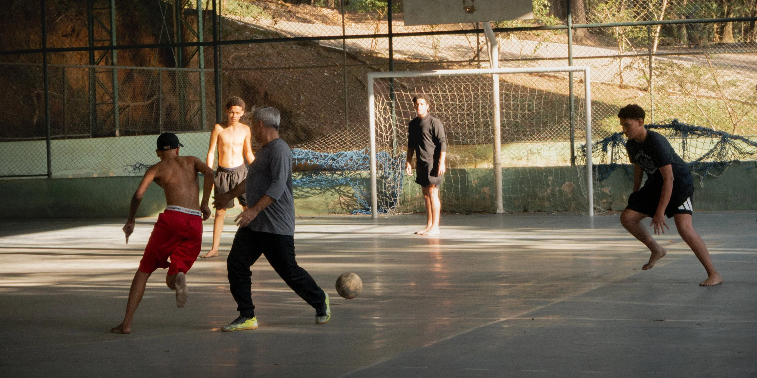 Jovens jogando futebol em quadra coberta, dois adolescentes de shorts, um rapaz mais velho, e uma goleira observando.