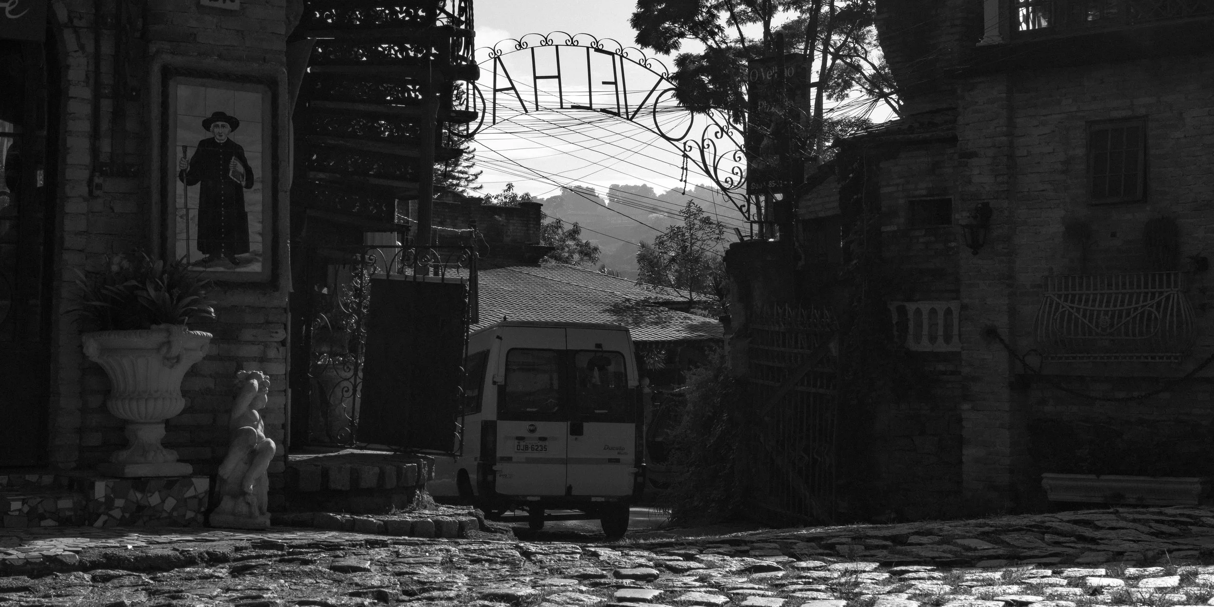 Rua de paralelepípedos com entrada fechada por uma cerca de ferro; ao lado, está uma escultura de uma mulher sentada, uma estátua de uma figura masculina com chapéu e uma planta em vaso. Ao fundo, há uma van branca estacionada e uma escada de ferro n
