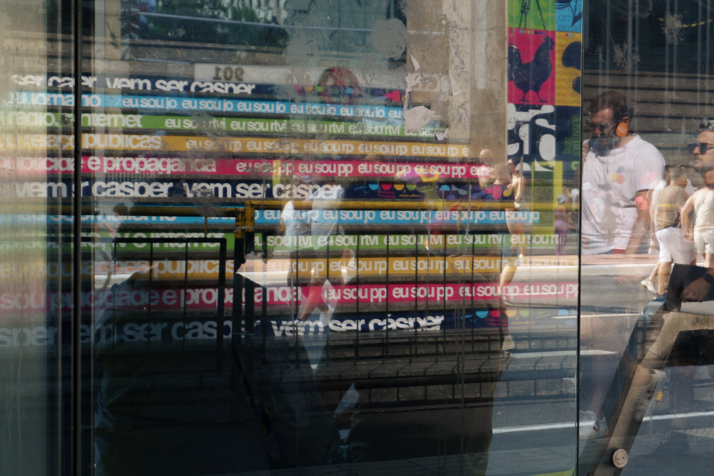 Reflexo de pessoas e faixas de protesto com textos em português refletindo em uma janela de vidro. Pessoas caminhando na rua ao fundo.