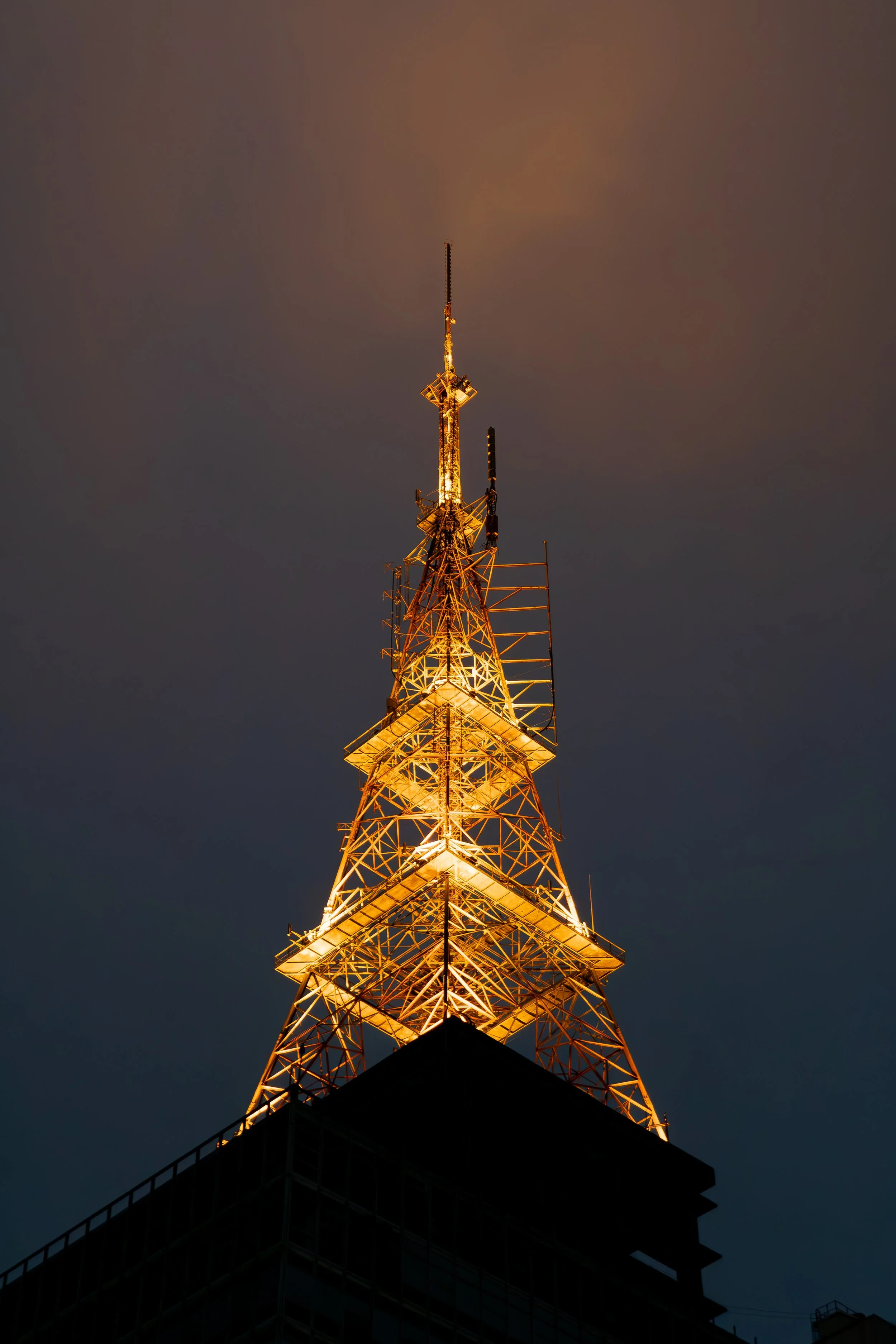 Torre de transmissão de rádio ou televisão iluminada à noite, com estrutura metálica destacada contra o céu escuro.