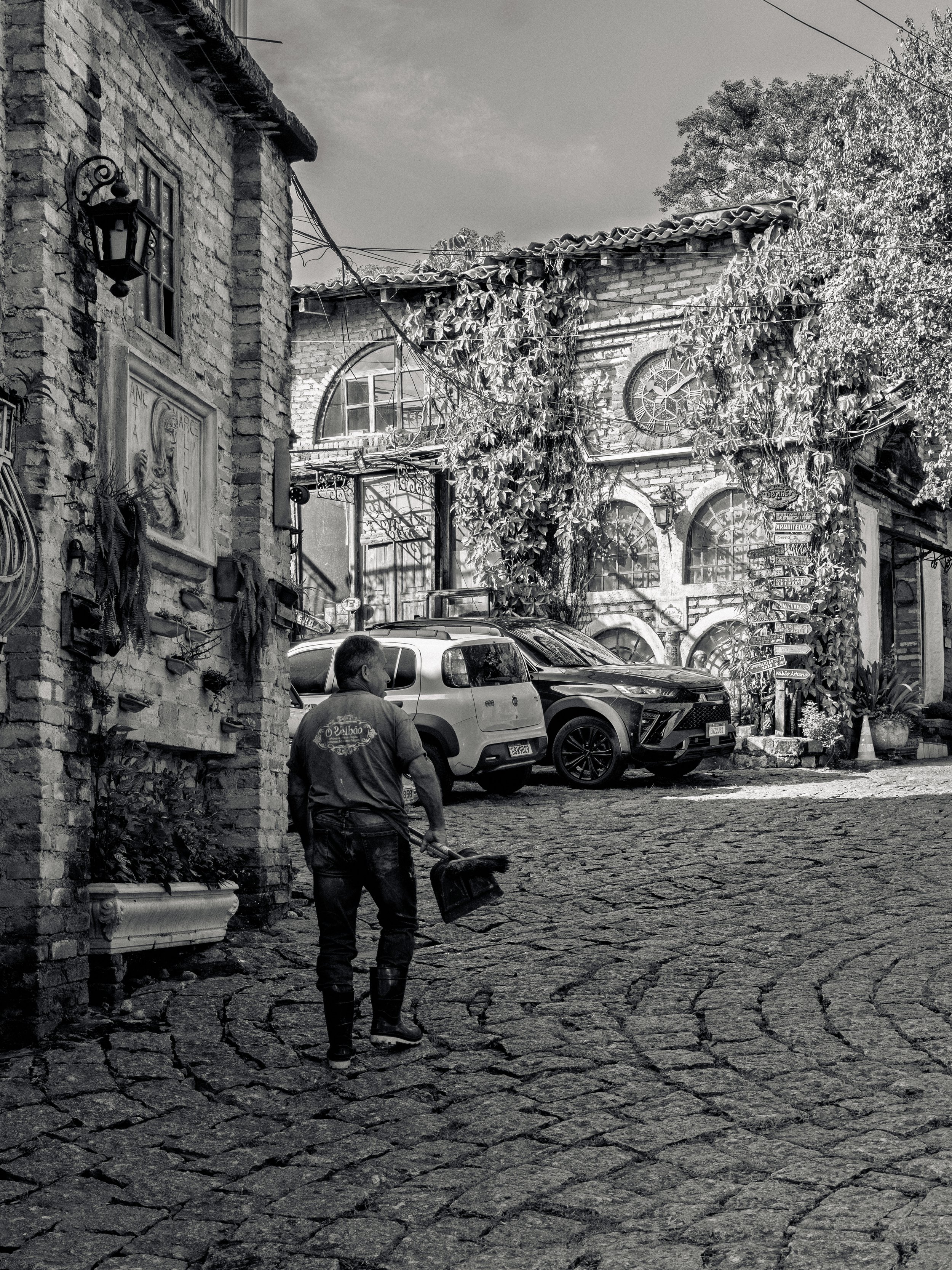 Pessoa varrendo a calçada de pedra em uma rua com casas de construção antiga, árvores e carros estacionados, em um ambiente ao ar livre.