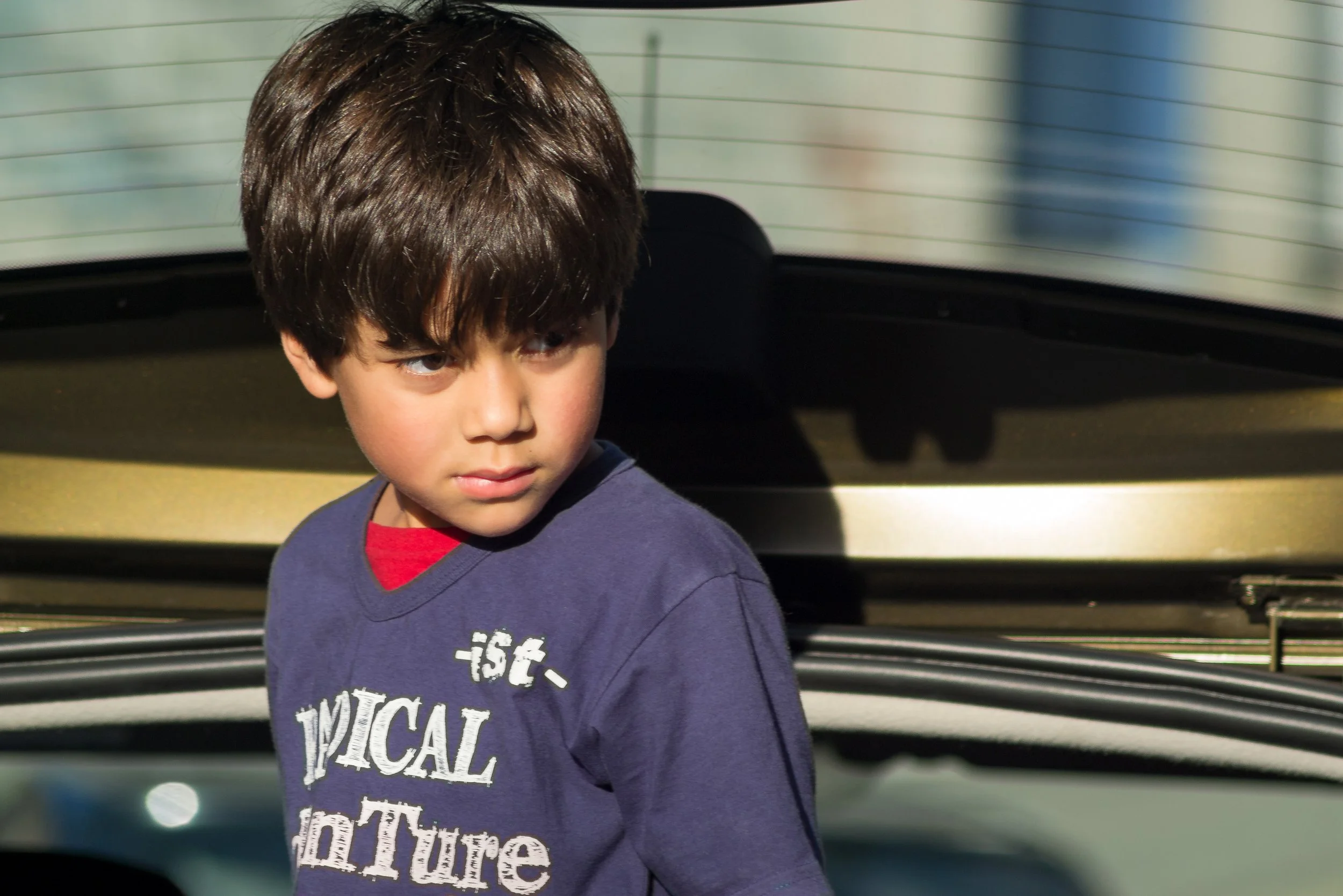 Menino com cabelo castanho e camiseta azul escuro com letras brancas, olhou para lado, com fundo de carro preto com vidro traseiro.