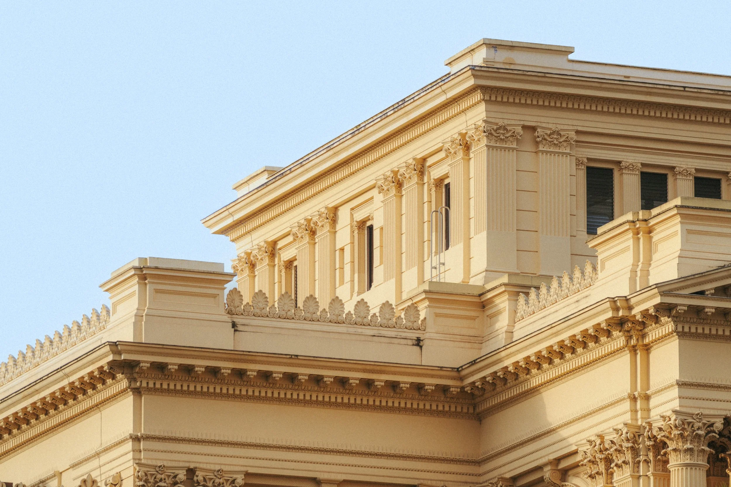 Imagem de uma construção clássica de cor creme com detalhes arquitetônicos elaborados, colunas e janelas, sob céu claro.