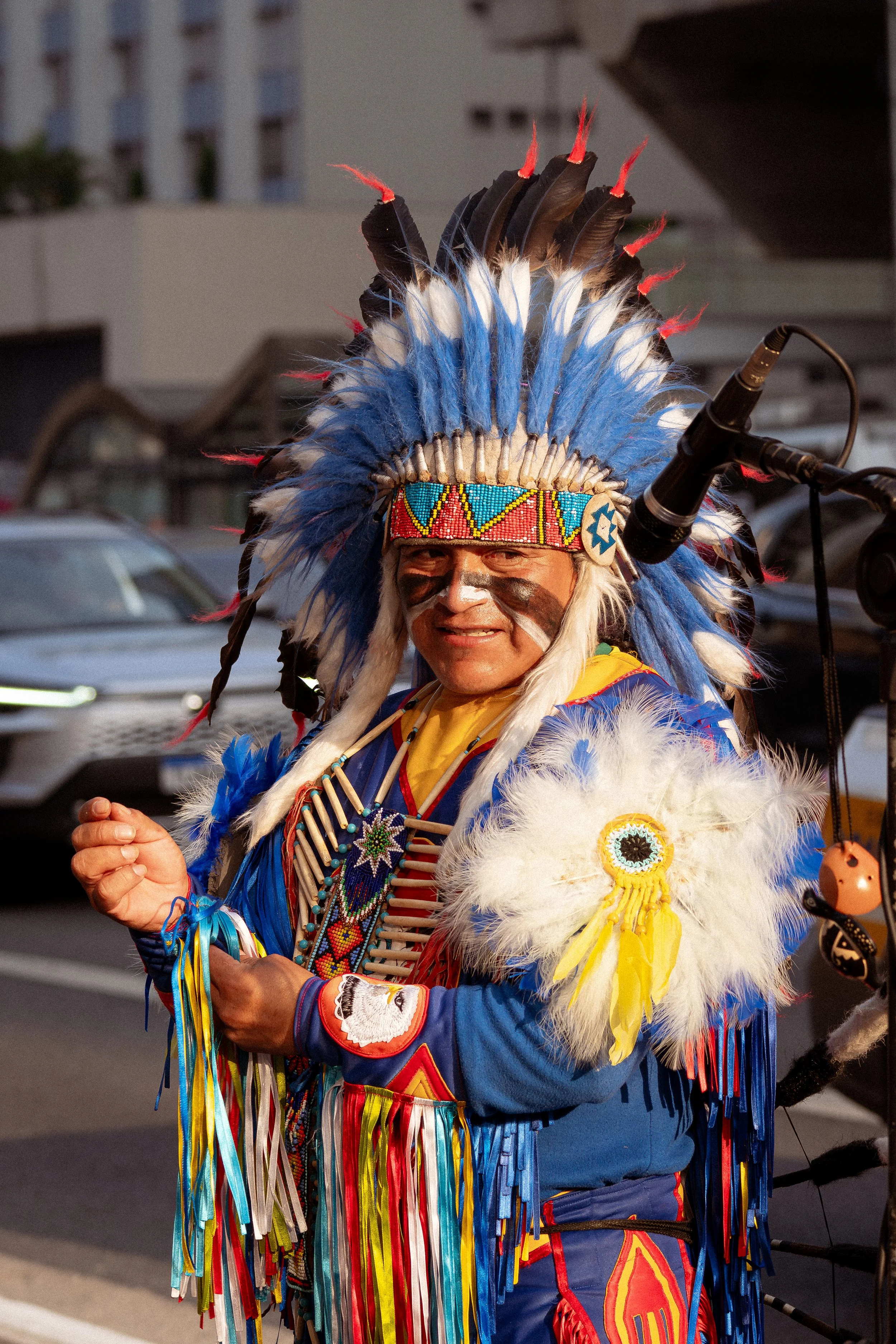 Homem de roupas tradicionais indígenas, com cocar e pintura facial, sorrindo durante uma celebração ao ar livre.