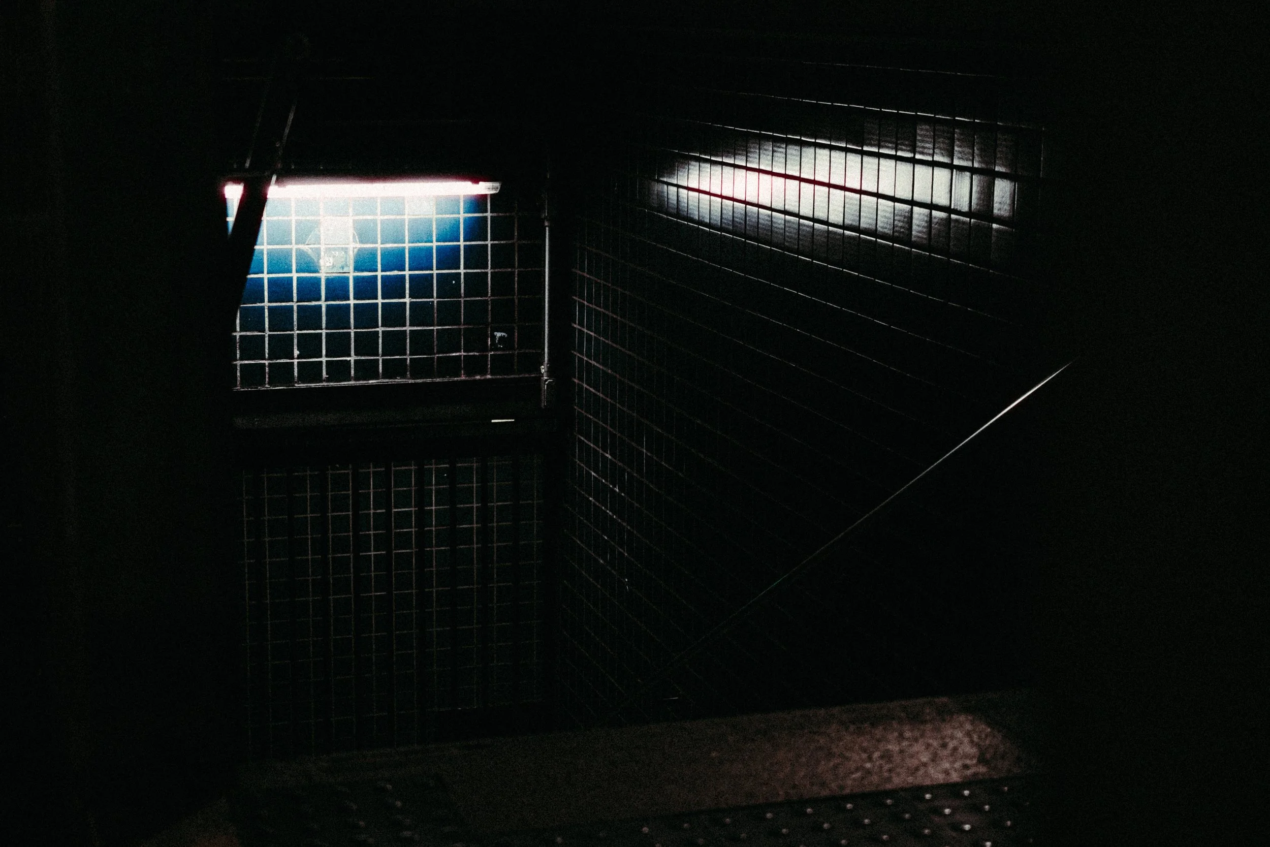 Imagem escura de uma escada com grades de ferro e uma pequena janela de onde entra luz.