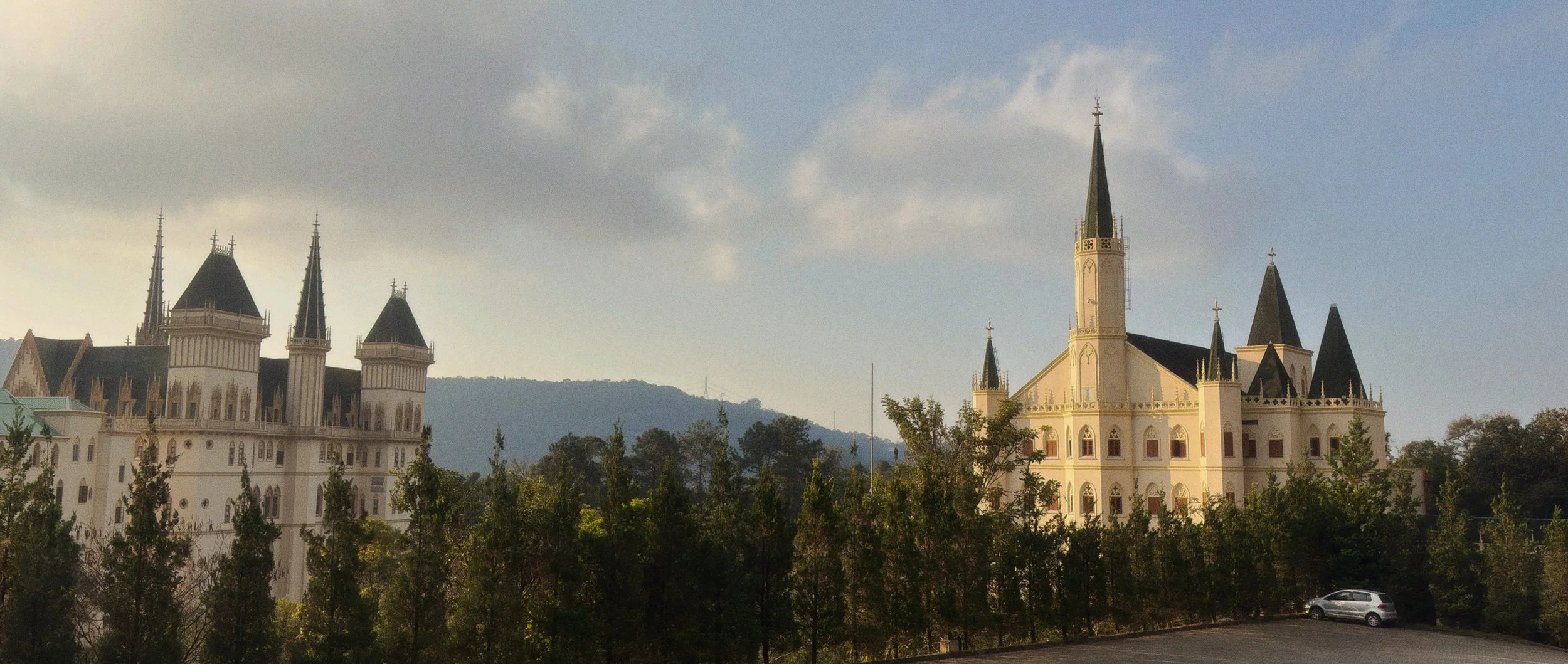 Castelo com torres pontiagudas e uma igreja com torre central, rodeados por árvores verdes e céu parcialmente nublado.