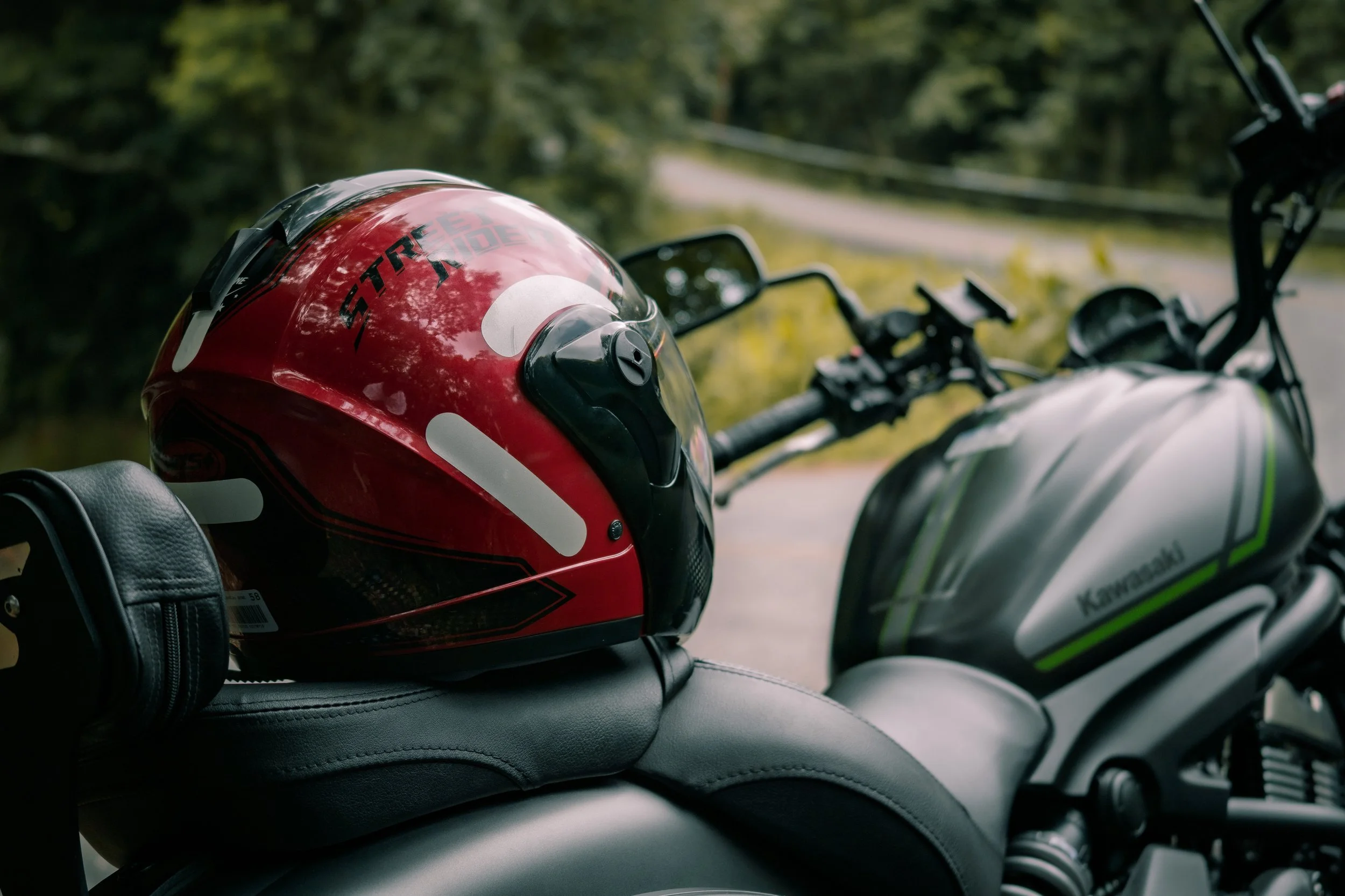 Capacete vermelho com detalhes em preto na parte superior da moto Kawasaki, com a marca escrita em preto, descansando na sela de uma moto preta e cinza com detalhes verdes, numa estrada ao lado de árvores verdes.