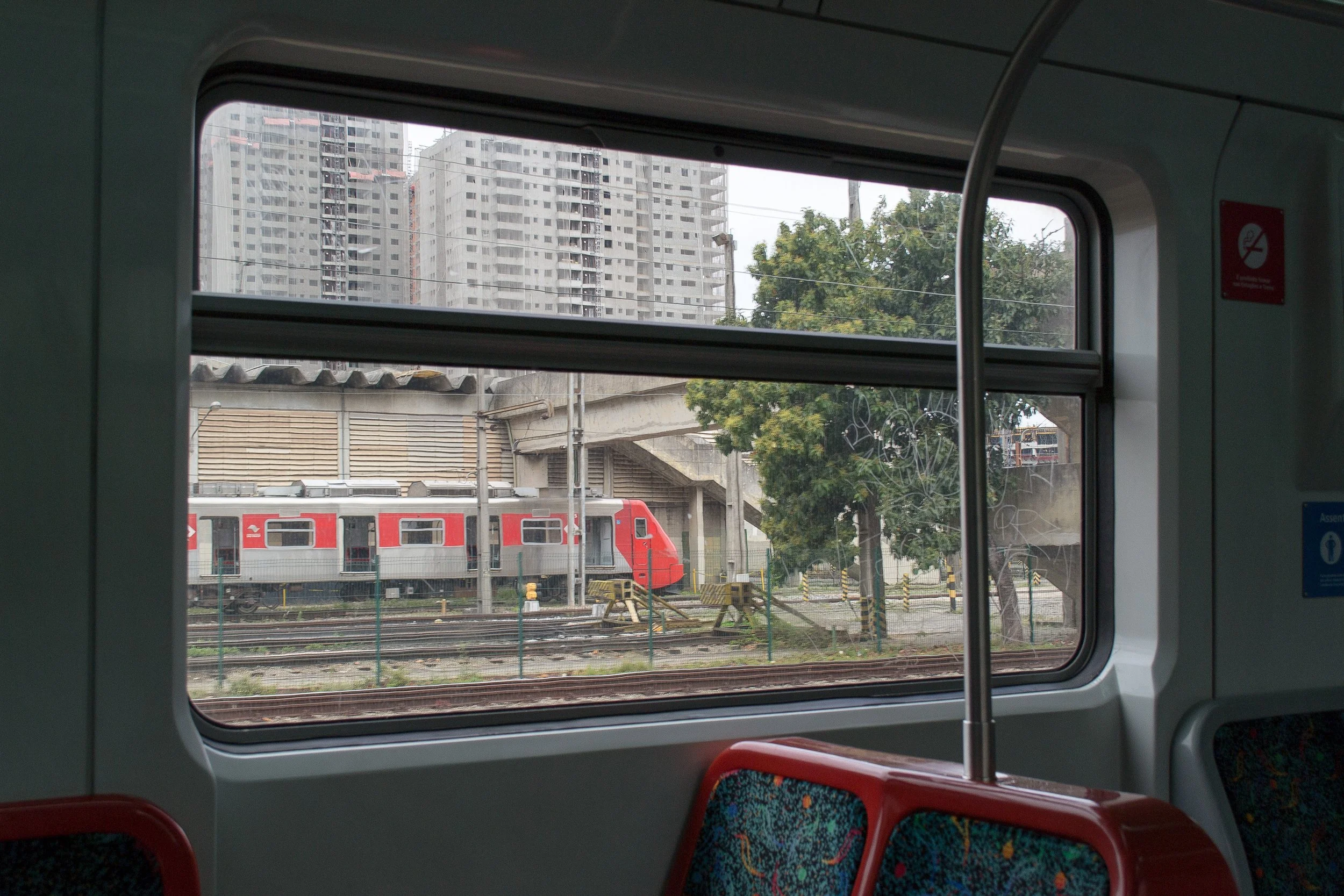 Janela de trem urbano com vista para ferrovias, prédio alto e árvores na cidade.