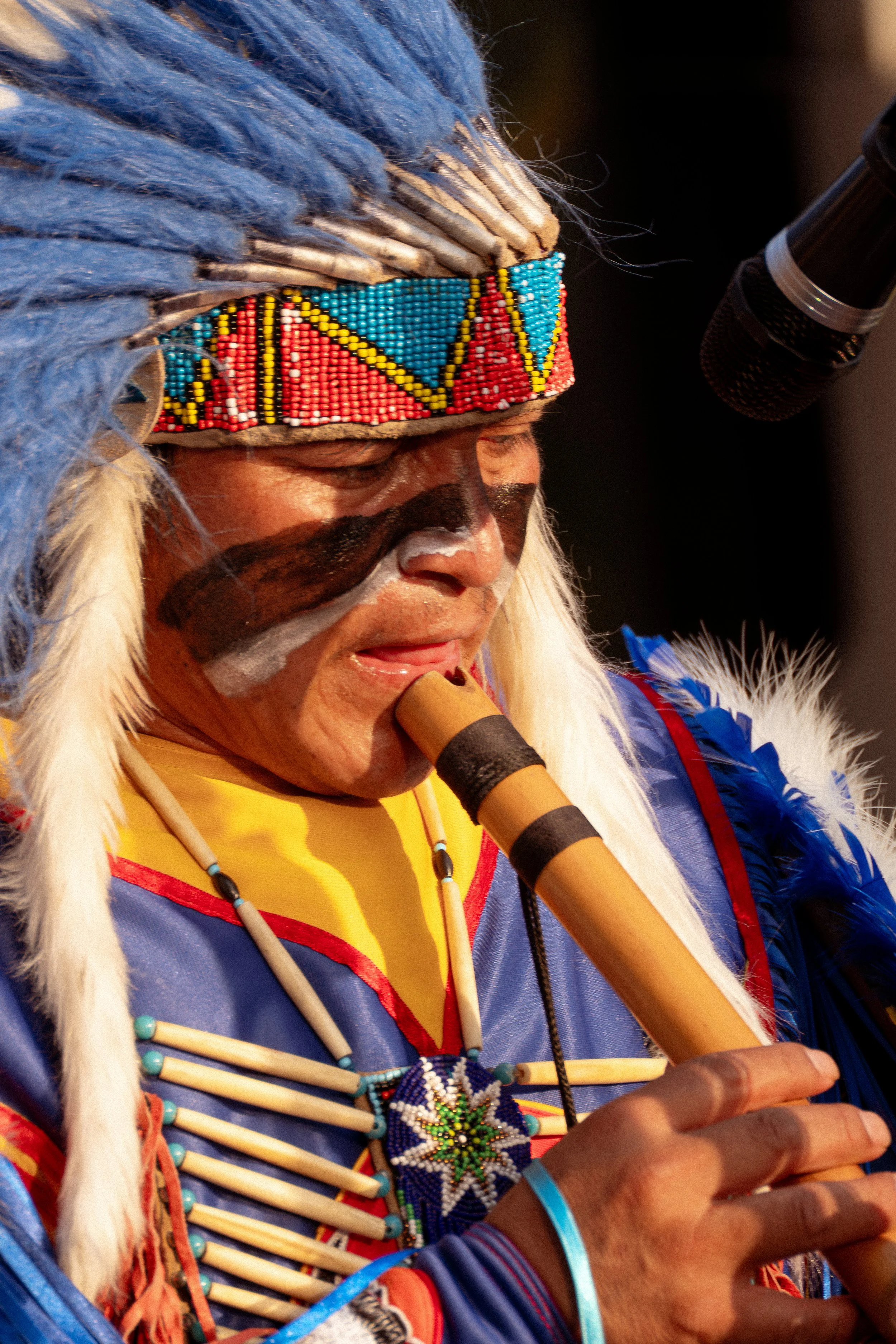 Homem indígena tocando uma flauta com vestimenta tradicional colorida, com detalhes em azul, vermelho, branco, usando adereços e maquiagem tribal, e uma pena no cabelo.