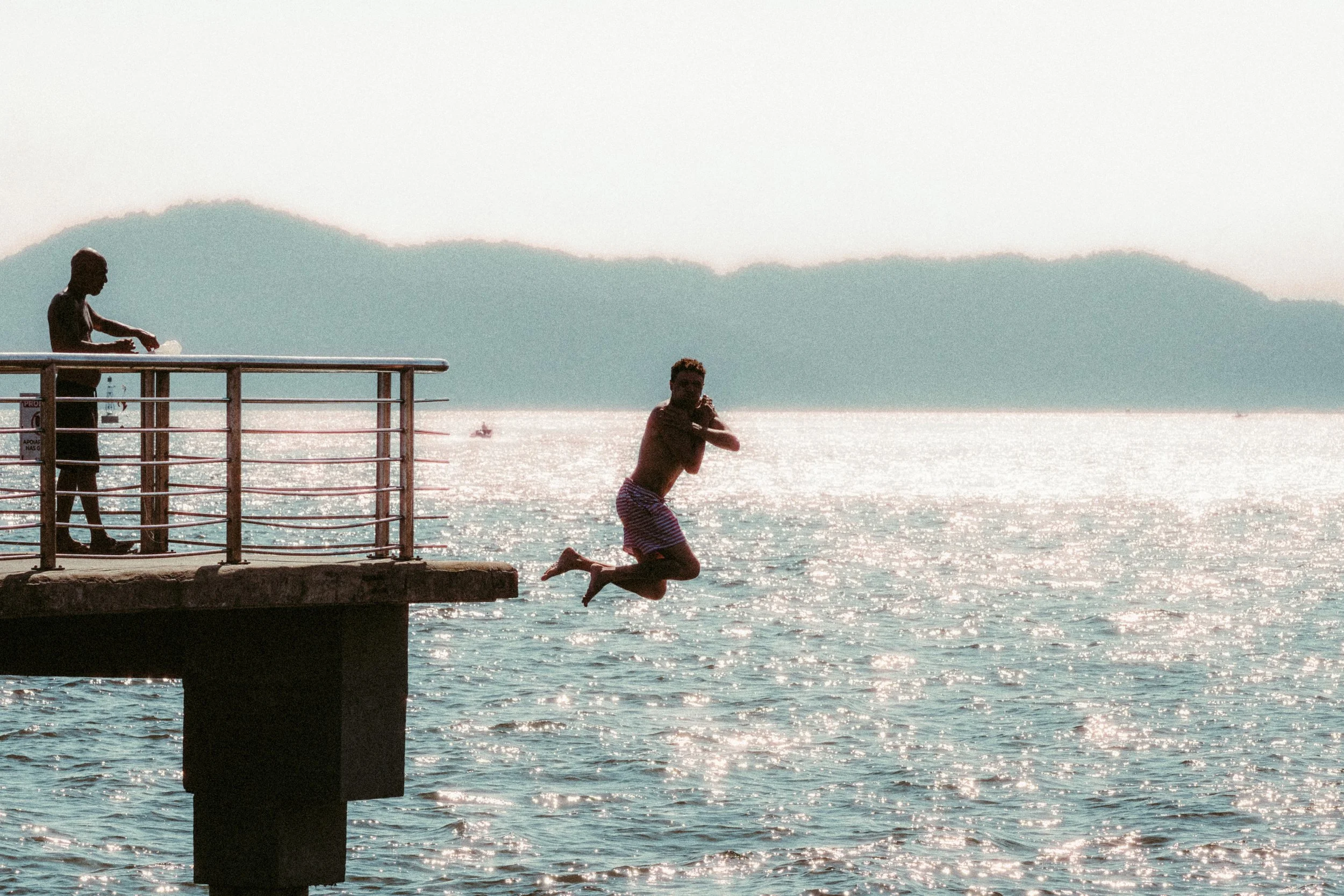 Homem pulando da ponte para o mar durante o pôr do sol, uma mulher observa na ponte, ao fundo há montanhas e o mar refletindo a luz do sol.