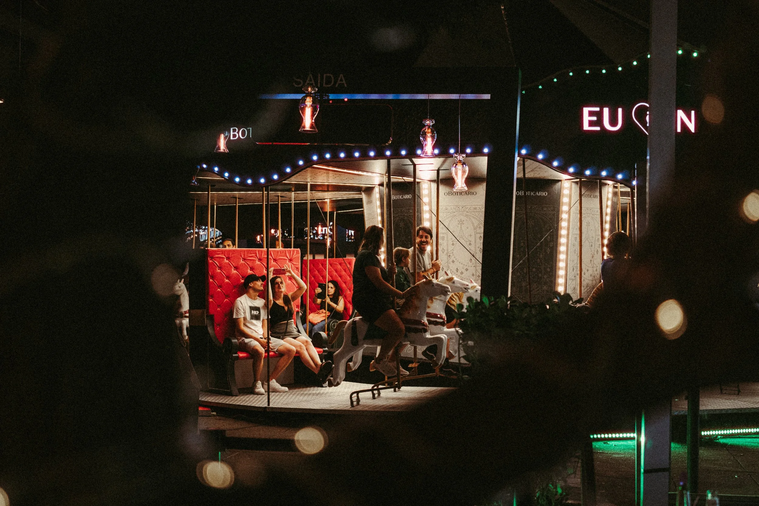 Diversas pessoas em um parque de diversões à noite, incluindo uma pessoa sorridente em um carrinho de cavalo de parque de diversões com fundo de luzes decorativas e sinais.