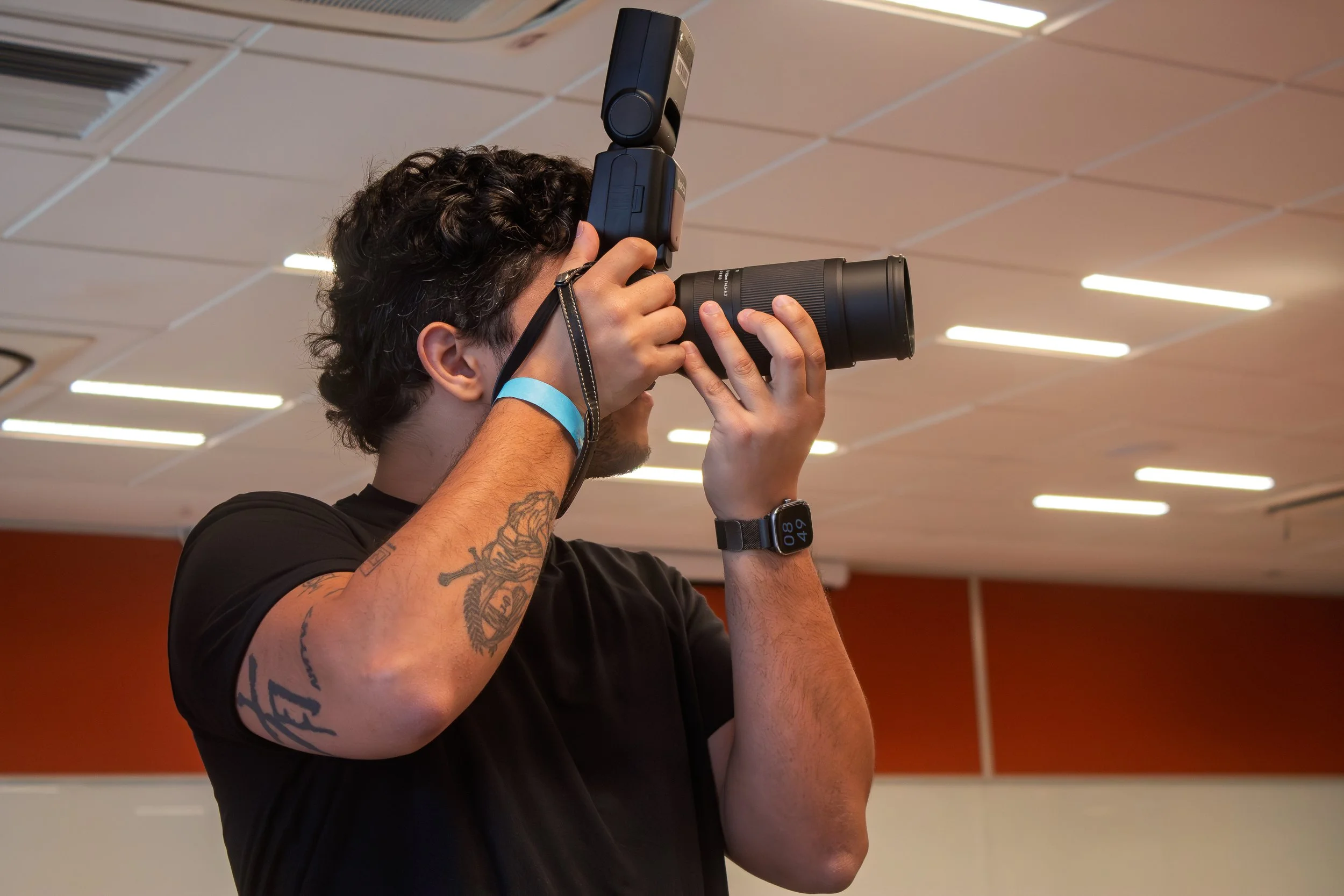 Pessoa segurando uma câmera fotográfica ao se posicionar para tirar uma foto, no interior de um ambiente com teto de gesso e iluminação suspensa.
