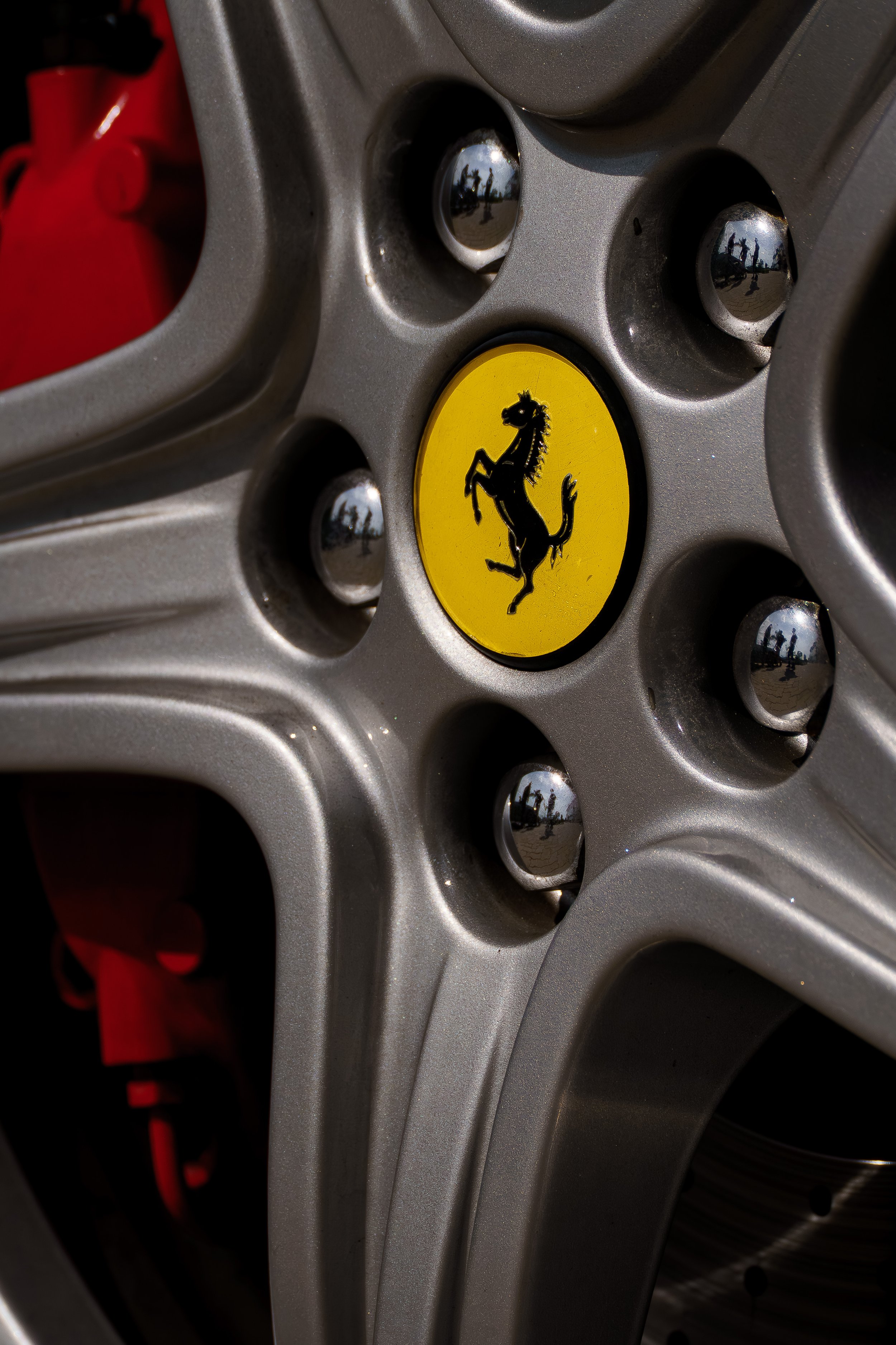 Roda de carro com logo da Ferrari no centro, mostrando detalhes de parafusos e reflexão da paisagem ao redor.