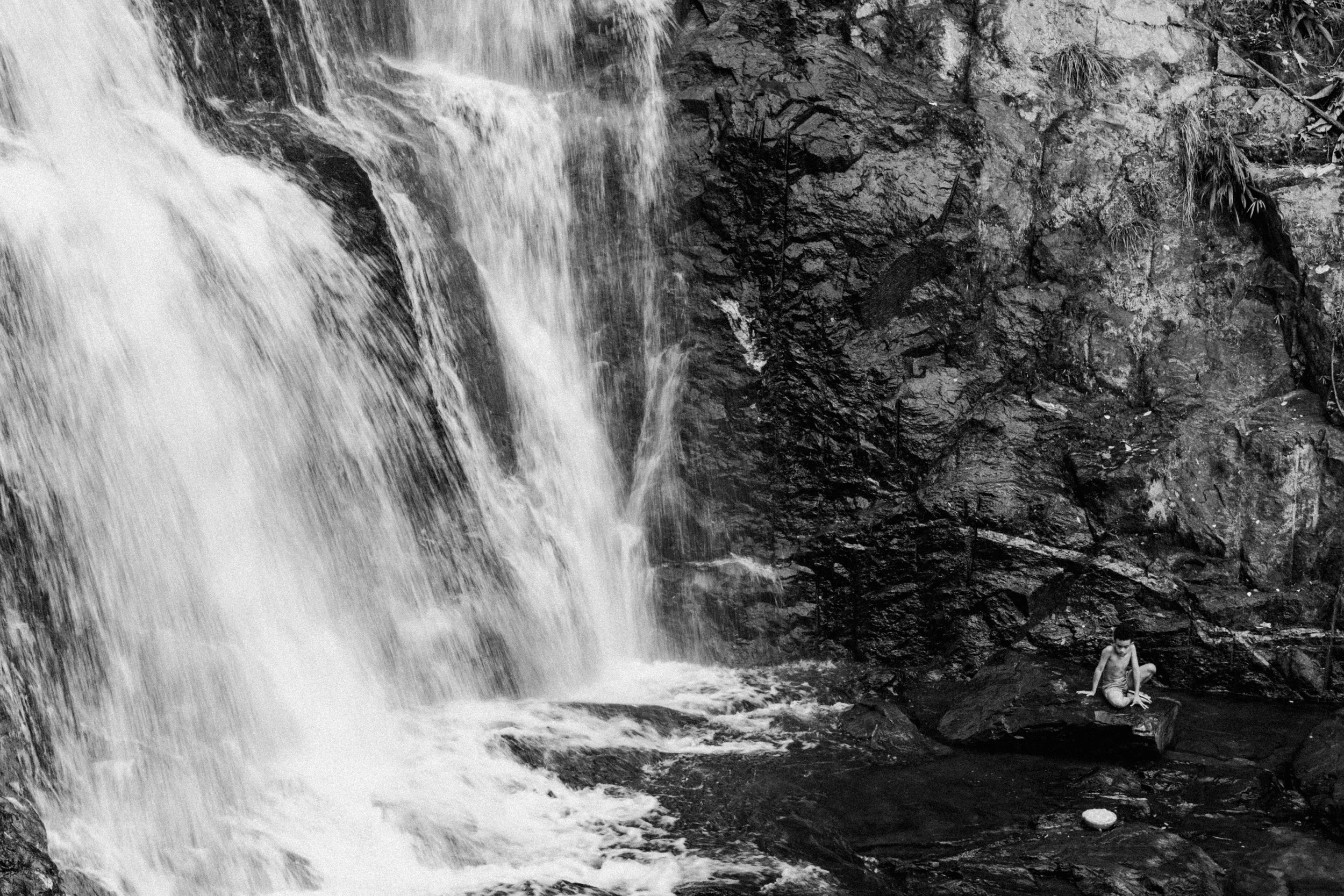 Criança sentado em uma pedra perto de uma cachoeira rochosa em ambiente natural, imagem em preto e branco.