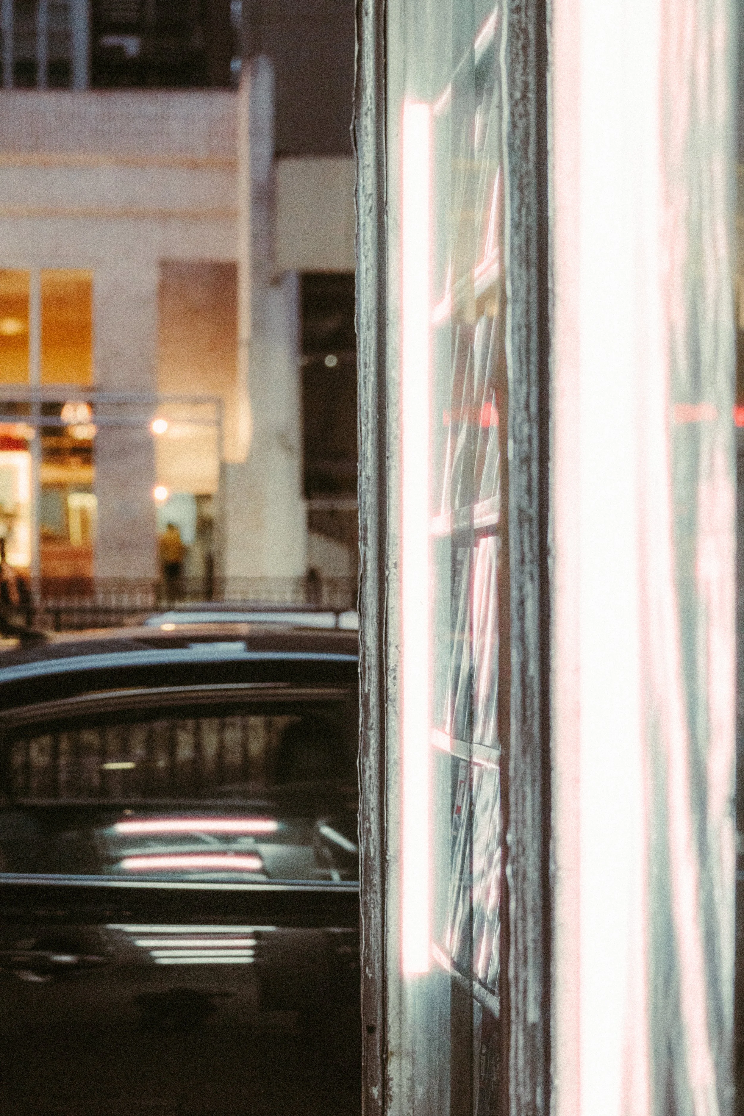 Foto da fachada de uma loja com janelas de vidro, refletindo luzes de néon, ao lado de uma rua com carro estacionado na cidade à noite.