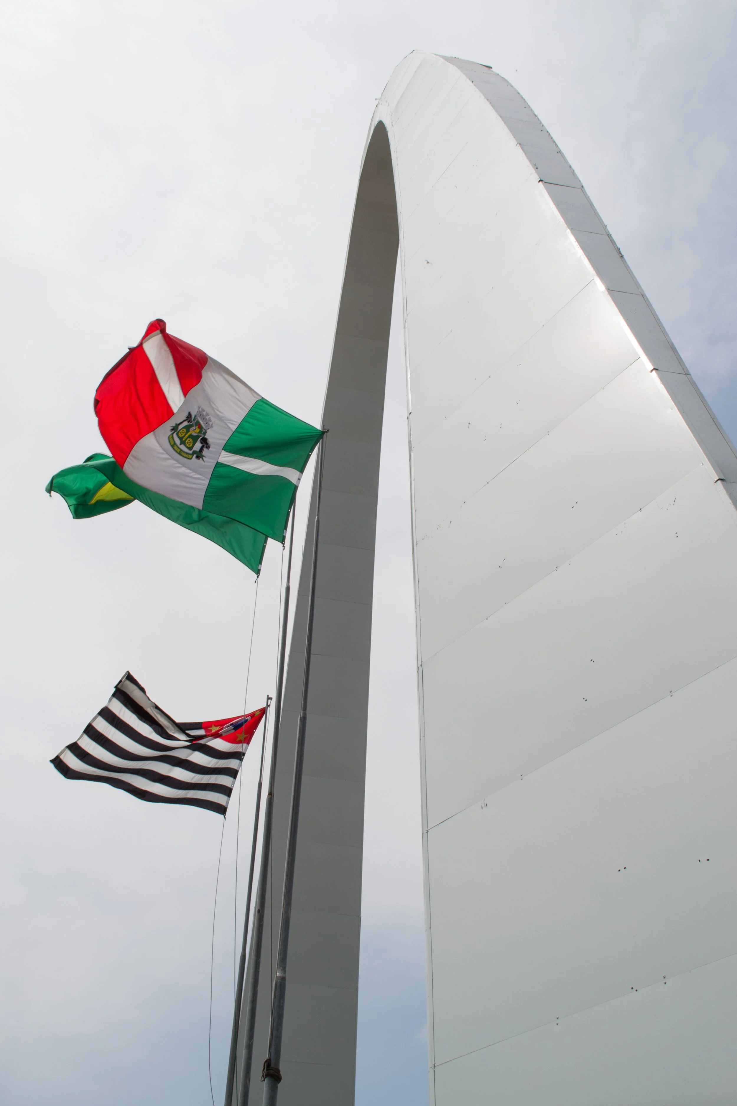 Bandeiras do Brasil, São Paulo e uma bandeira com listras horizontais pretas e brancas, voando ao lado do arco da Guanabara contra céu nublado.