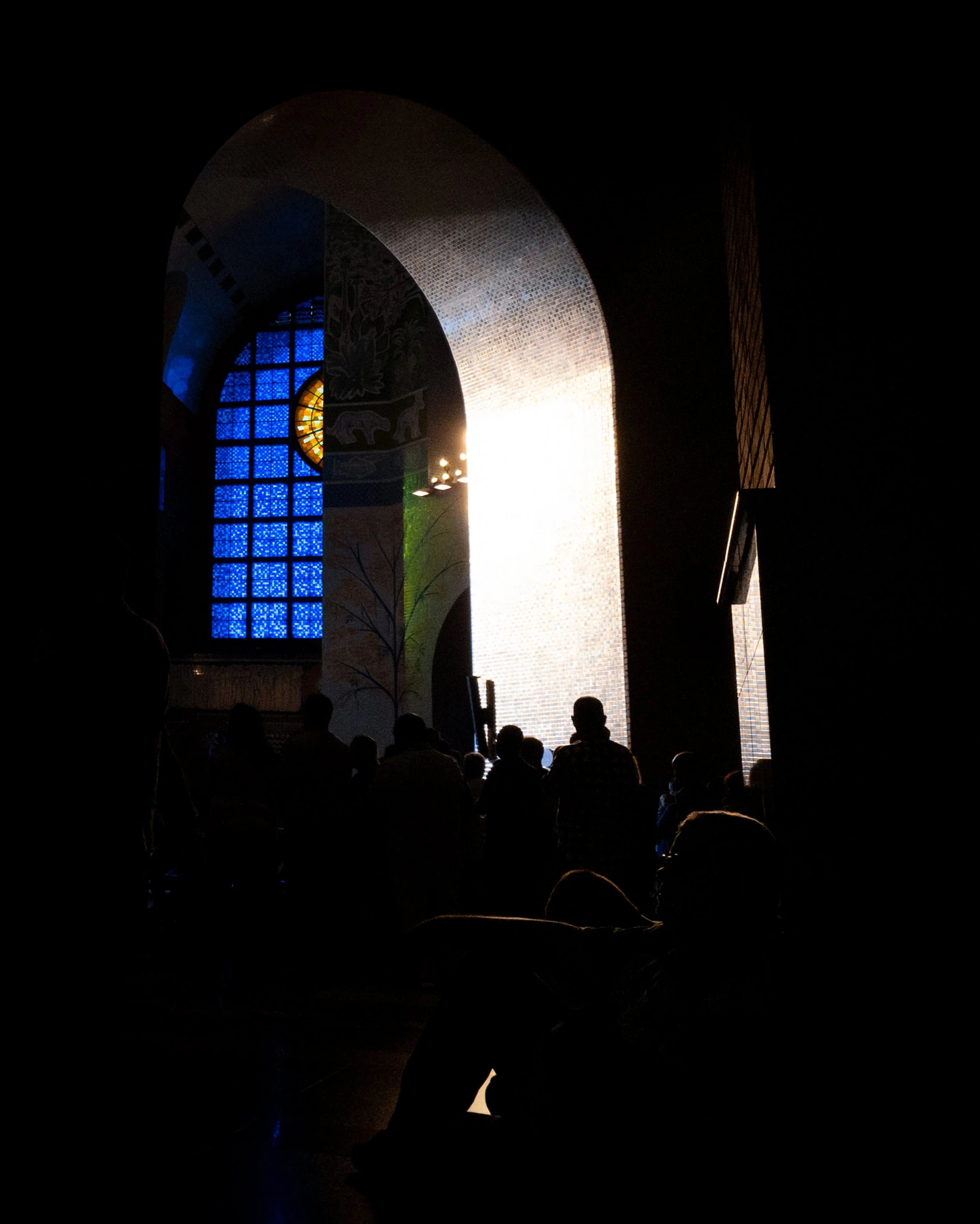 Interior de igreja com vitral azul e luz entrando, silhuetas de pessoas sentadas e de pé.