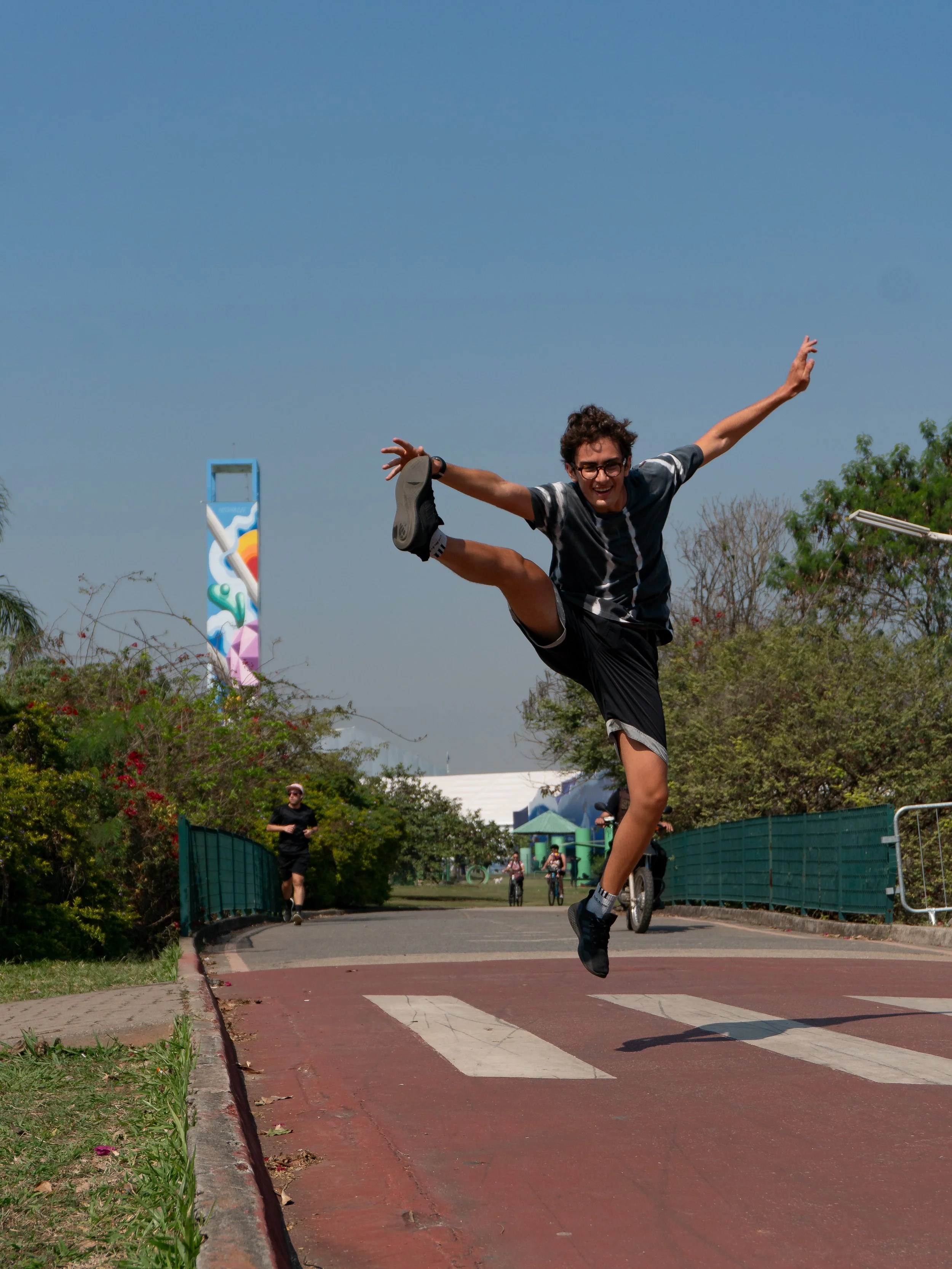 Pessoa fazendo uma acrobacia no parque, pulando no ar com expressão de alegria, outros ciclistas ao fundo, árvores e céu azul claro.