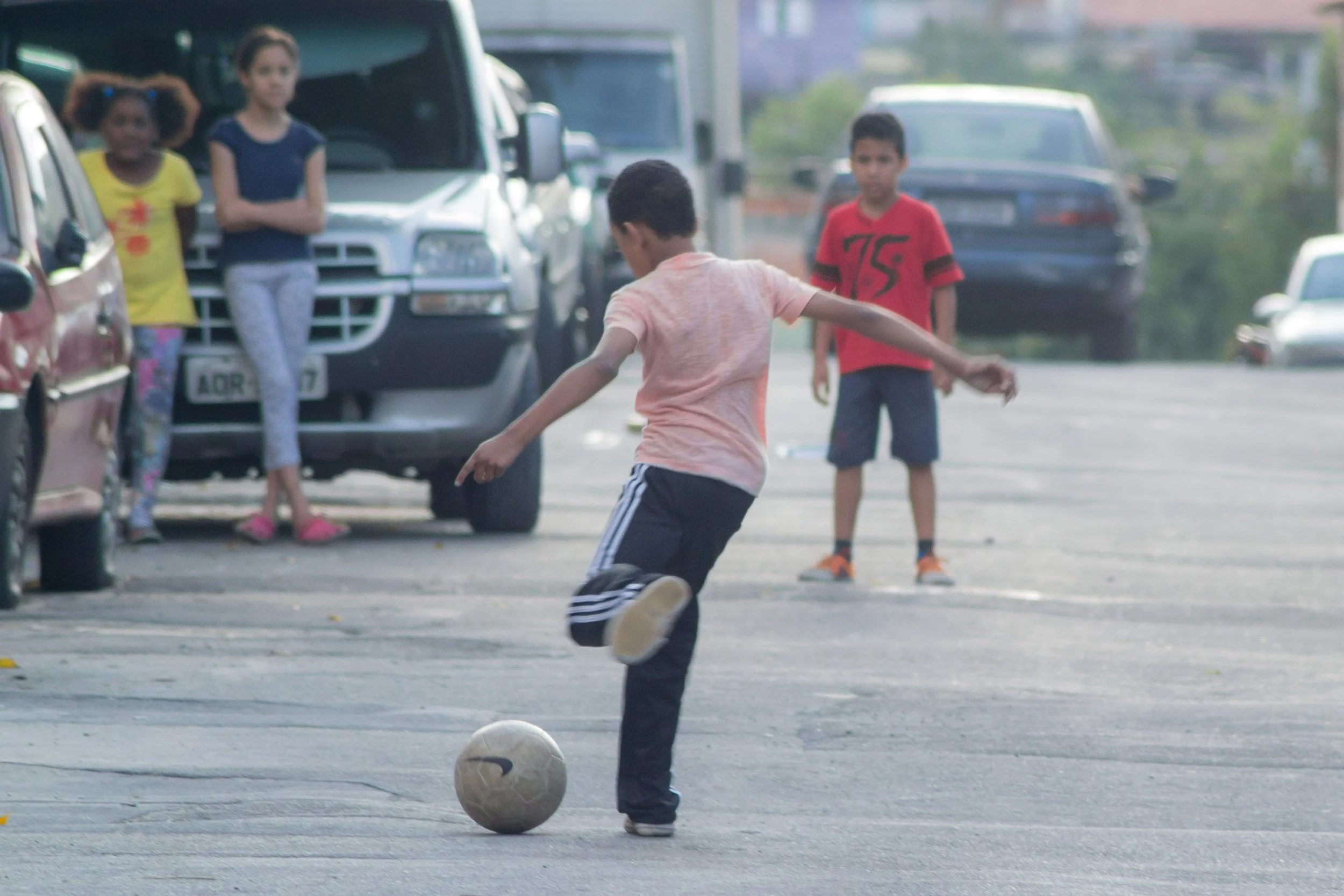 Crianças jogando futebol na rua, com alguns carros estacionados ao fundo.