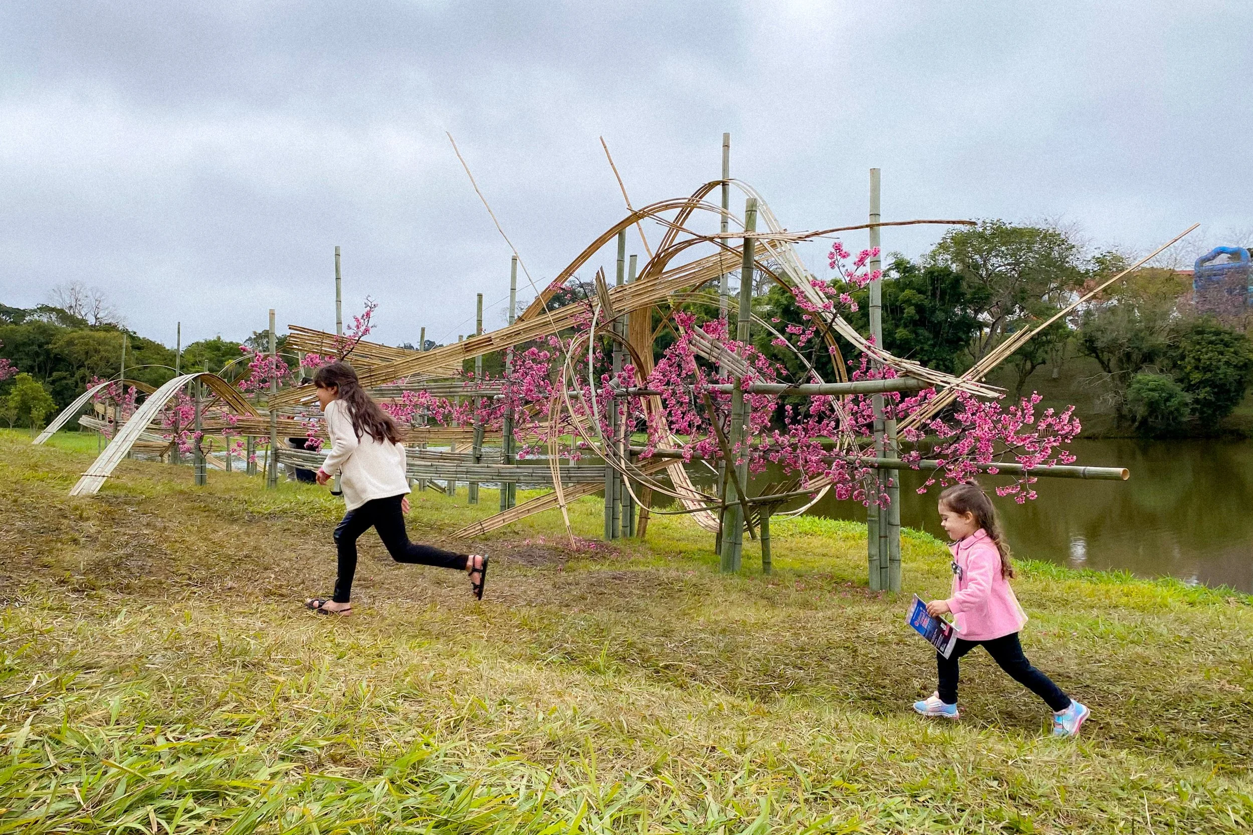 Duas crianças brincando ao ar livre perto de uma estrutura de bambu decorada com flores rosas, ao lado de um rio, sob céu nublado.