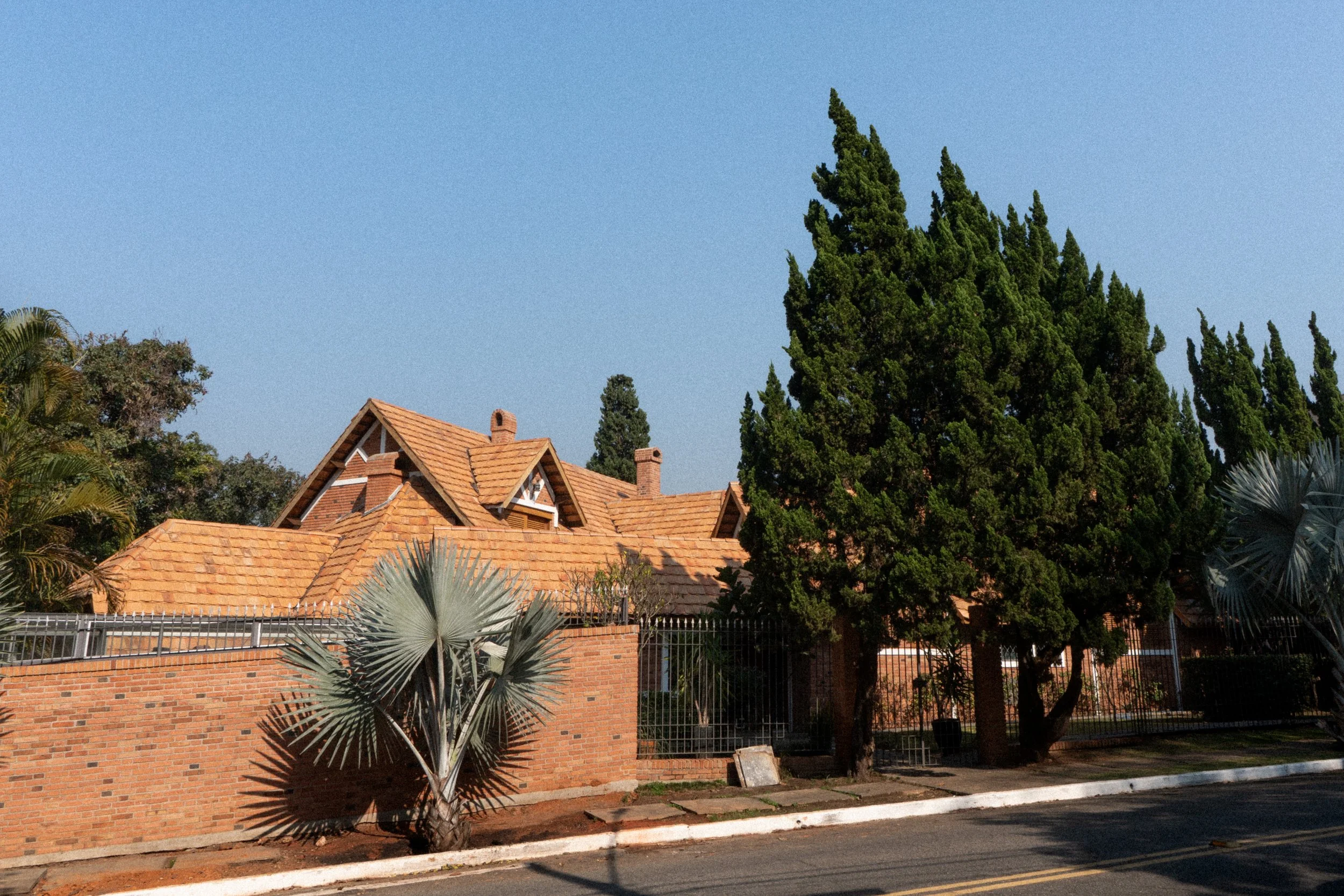 Casa com telhado de losango, muro de tijolos e árvores em volta, sob céu azul.