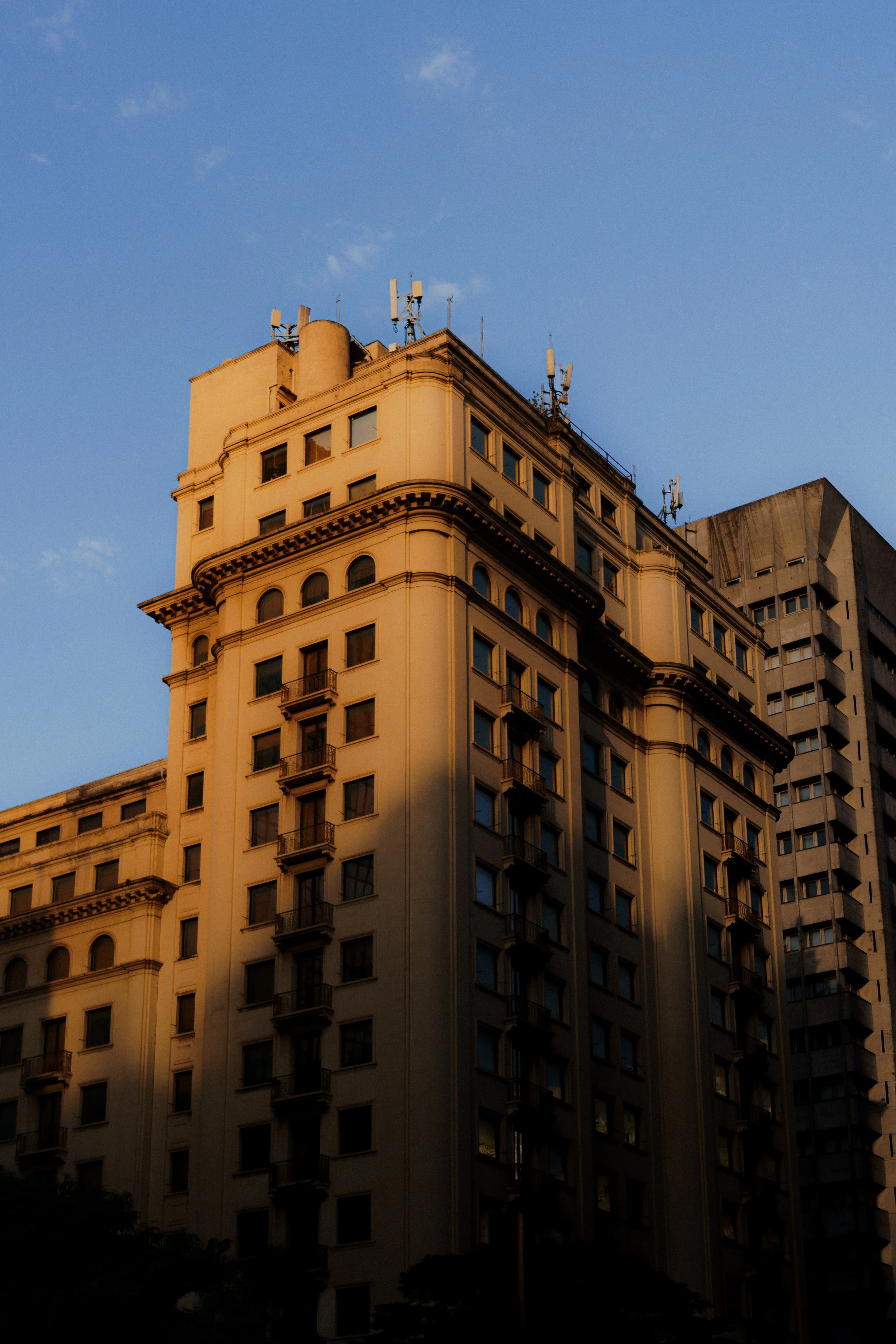 Prédio antigo de cor amarela com vários andares, janelas pequenas e sacadas, sob céu azul ao entardecer.