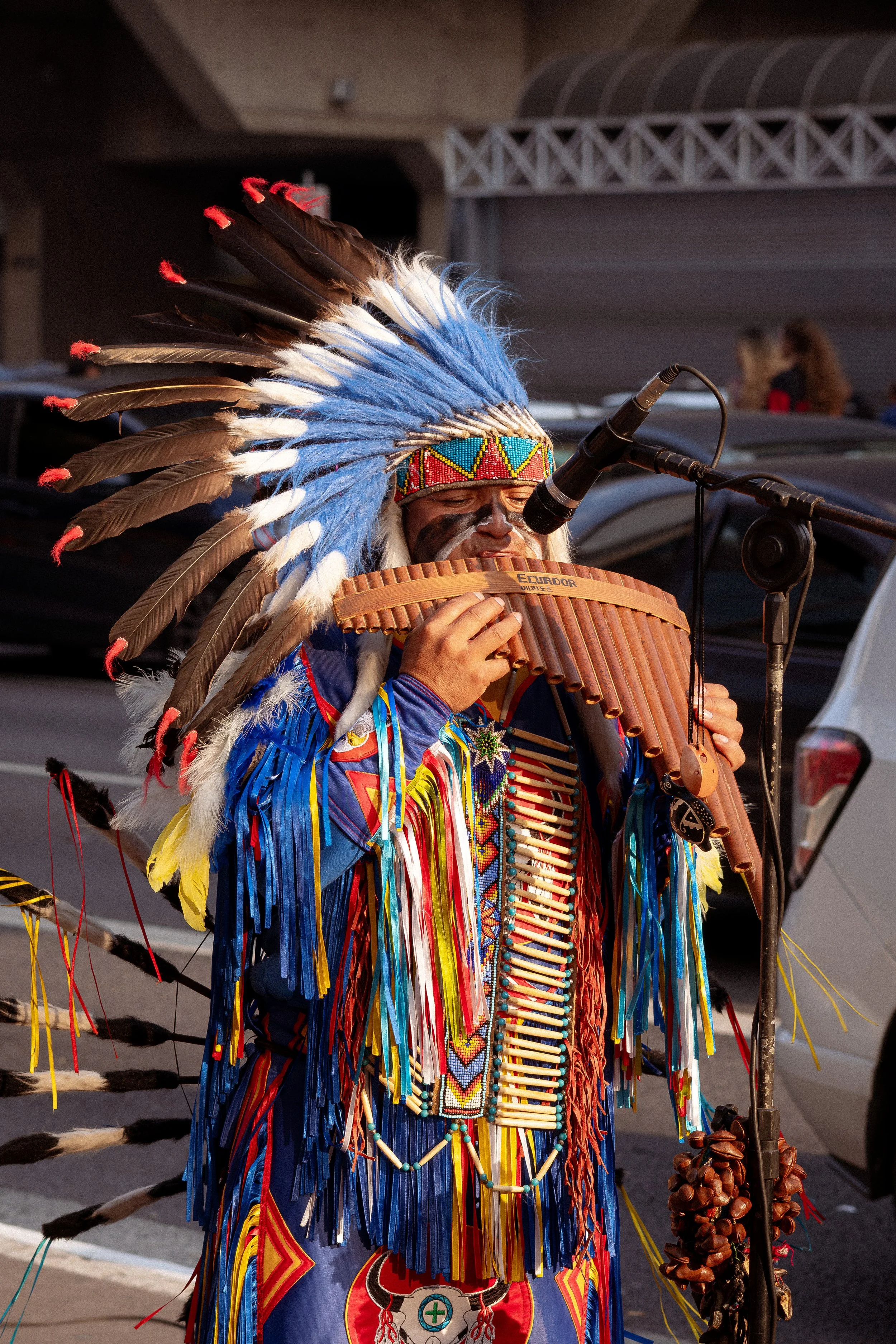 Homem indígena com vestimenta tradicional colorida, tocando uma flauta e cantando, usando um adereço de pena na cabeça durante uma apresentação ao ar livre.