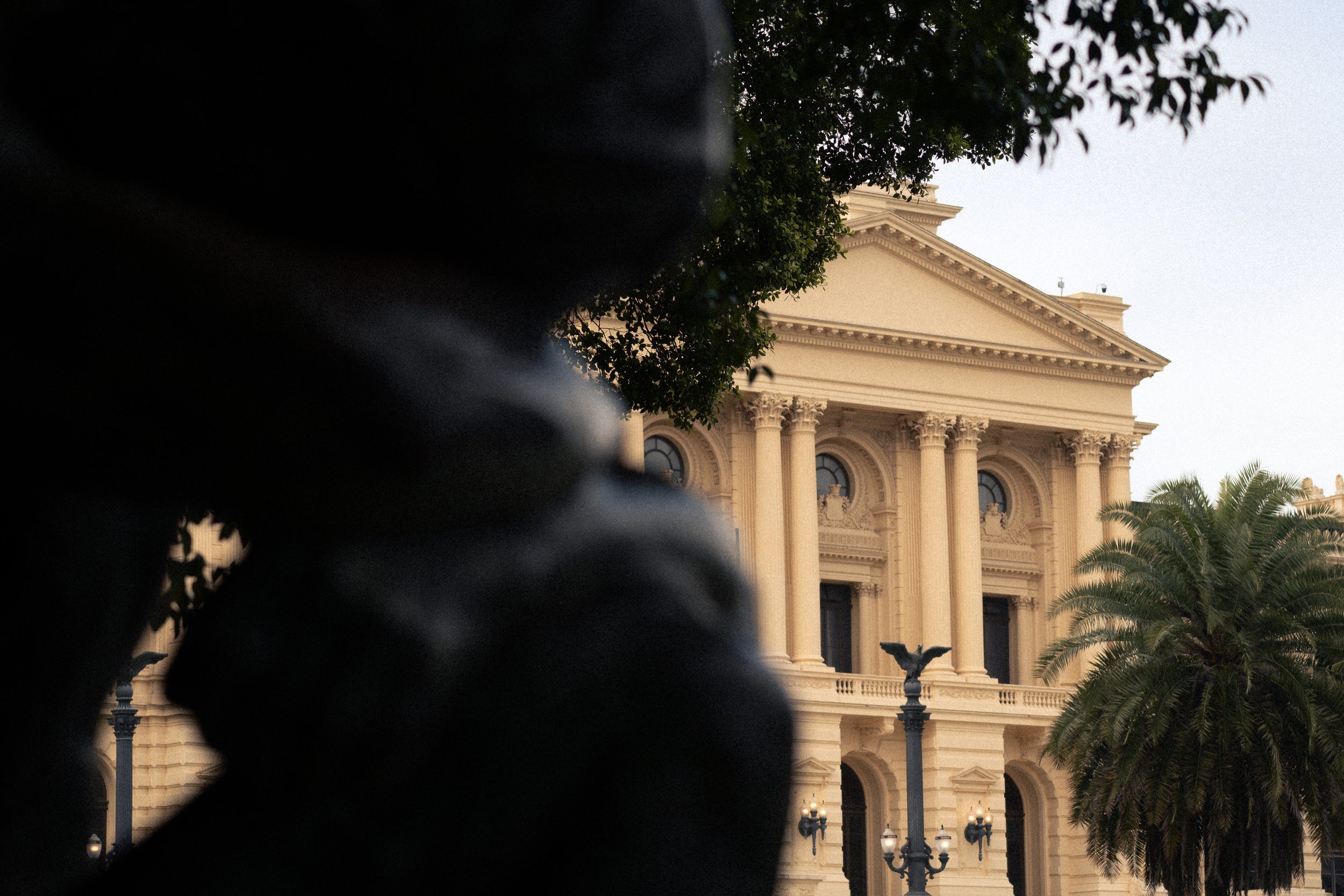 Edifício clássico com colunas, árvores e estátuas, parcialmente escondido por sombra ou objeto preto na frente.