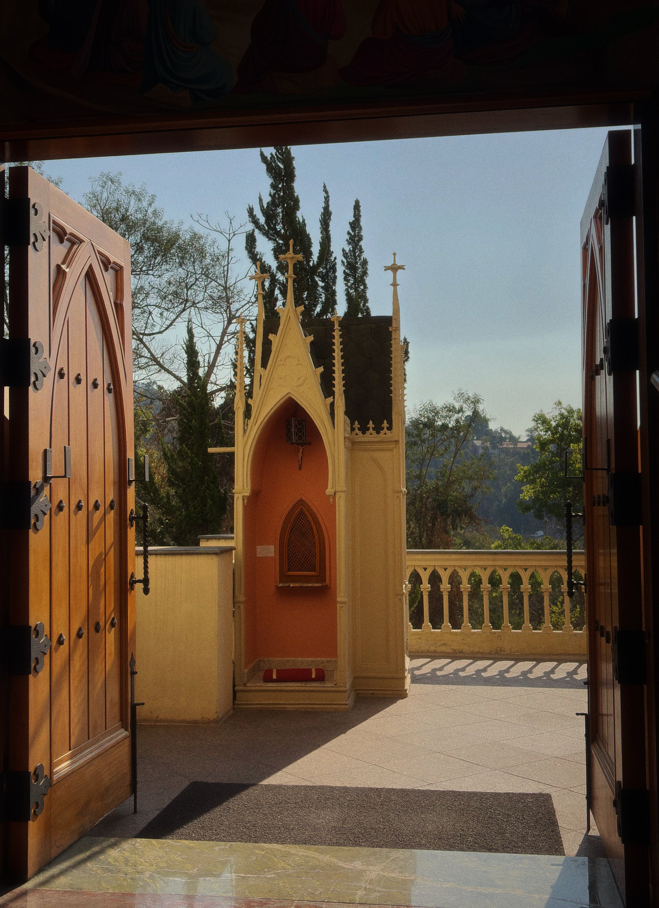 Vista do interior de uma capela ou igreja, com portas de madeira abertas revelando um altar ao ar livre, com árvores e céu ao fundo.