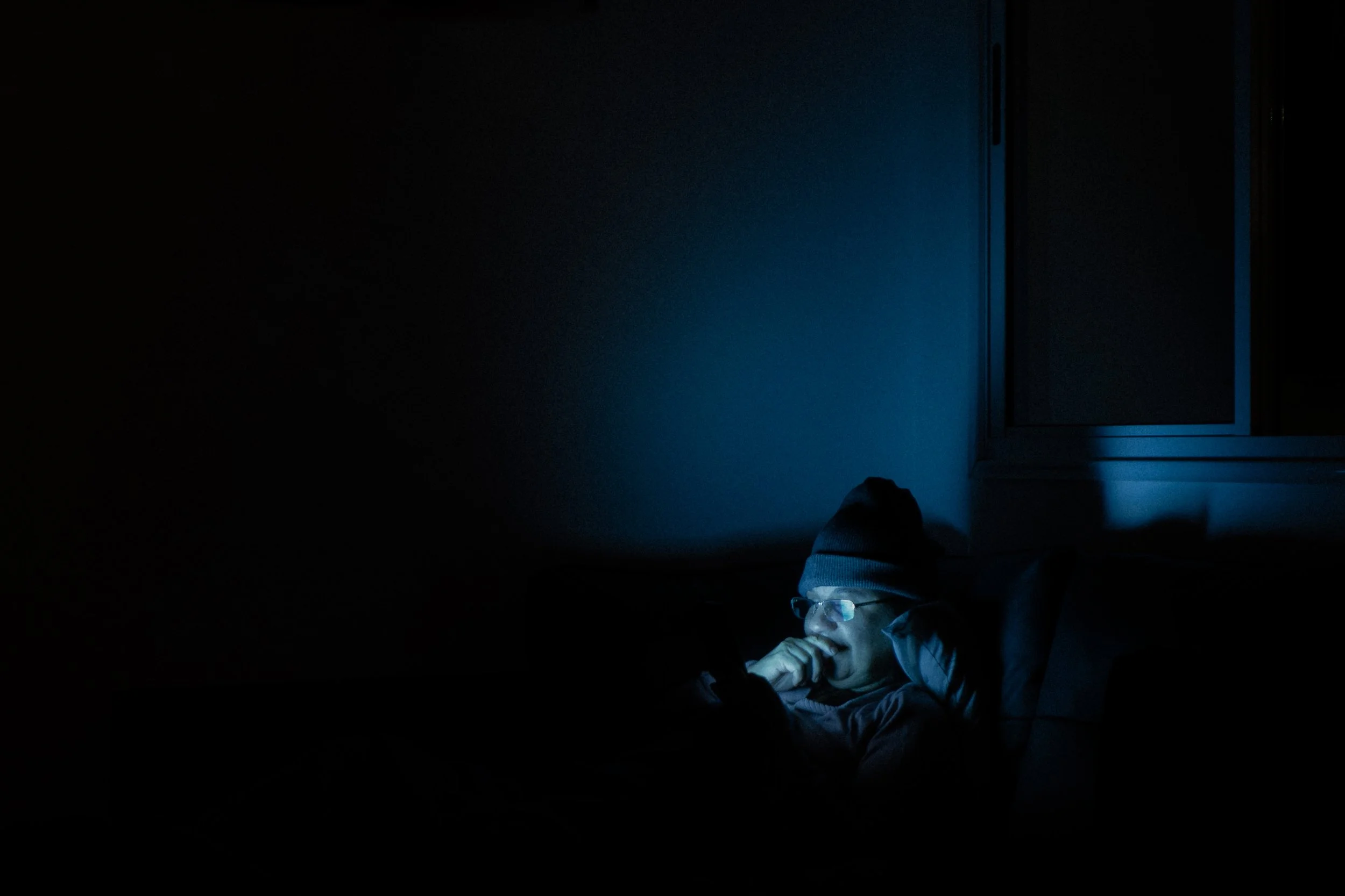 Pessoa usando óculos e gorro, deitado na cama, iluminada pela luz azul de uma tela, no escuro de um quarto à noite.