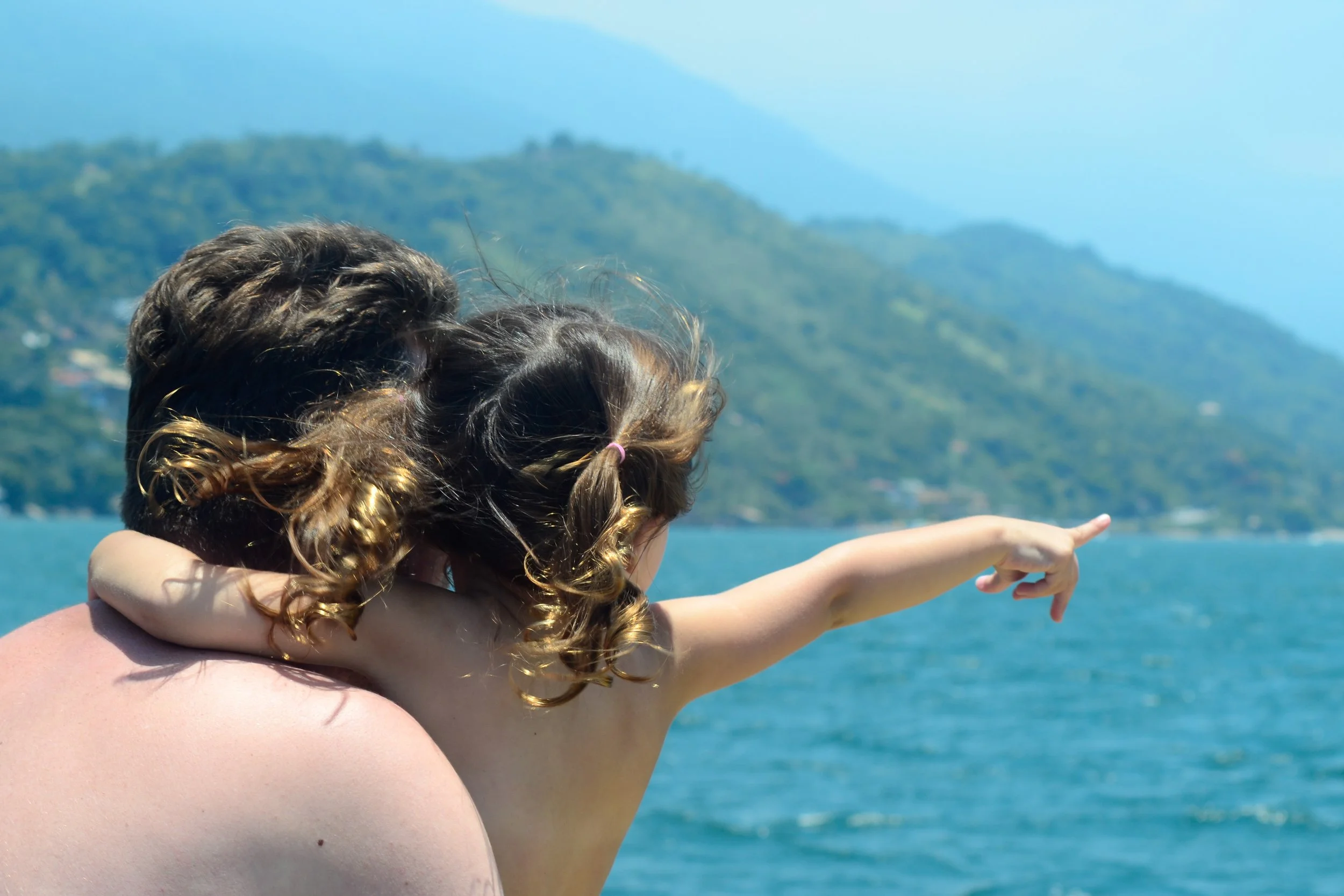 Um adulto segurando uma criança que está apontando para o mar, com uma paisagem de montanhas ao fundo.