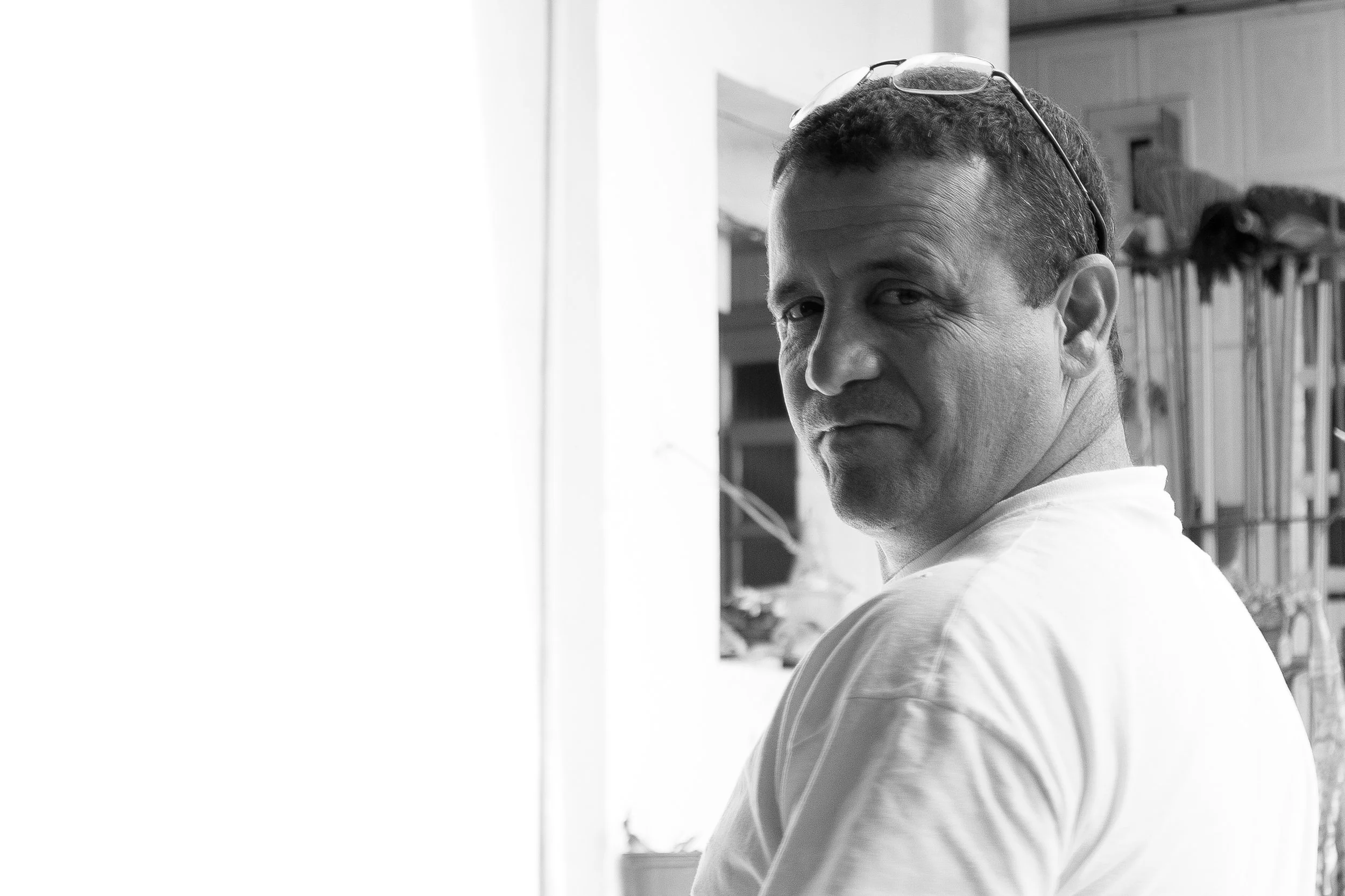 Homem com óculos na cabeça, virado de costas, olhando para trás. Fundo com prateleiras de utensílios e objetos.