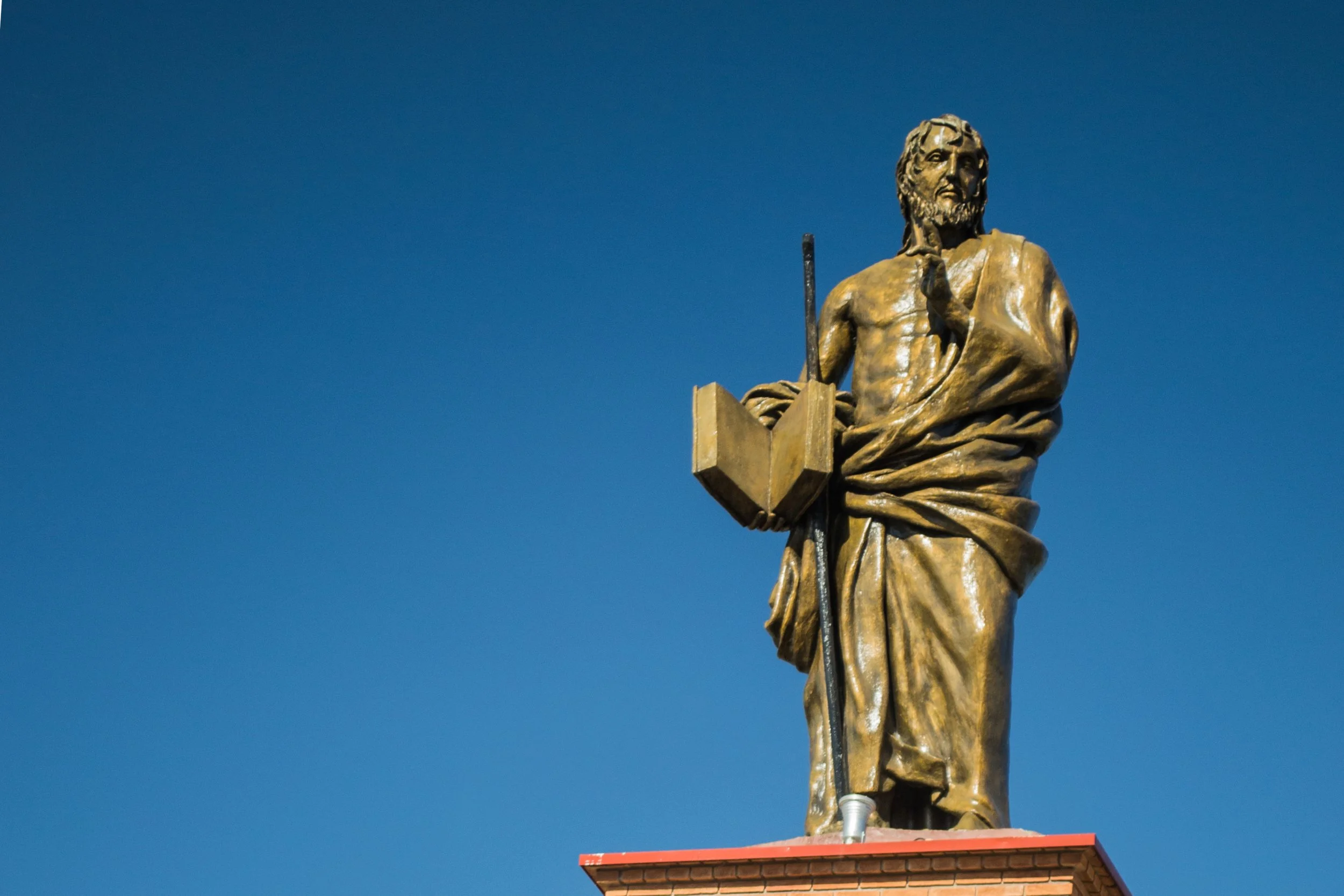 Escultura de uma figura masculina antiga, de aparência sacerdotal, com barba e cabelo longo, segurando um livro aberto na mão esquerda e com uma túnica drapeada, colocada sobre uma base de tijolos, contra um céu azul claro.