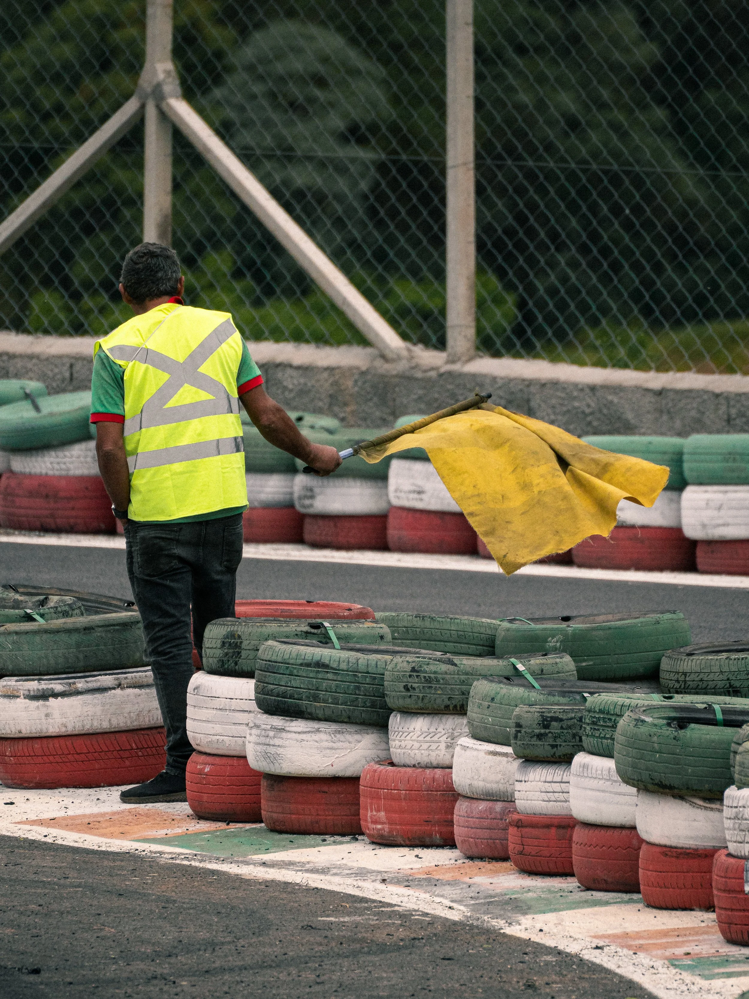 Pessoa com colete de segurança amarelo limpando faixas de pista de corrida com bandeira amarela, ao lado de pneus empilhados ao lado da pista.