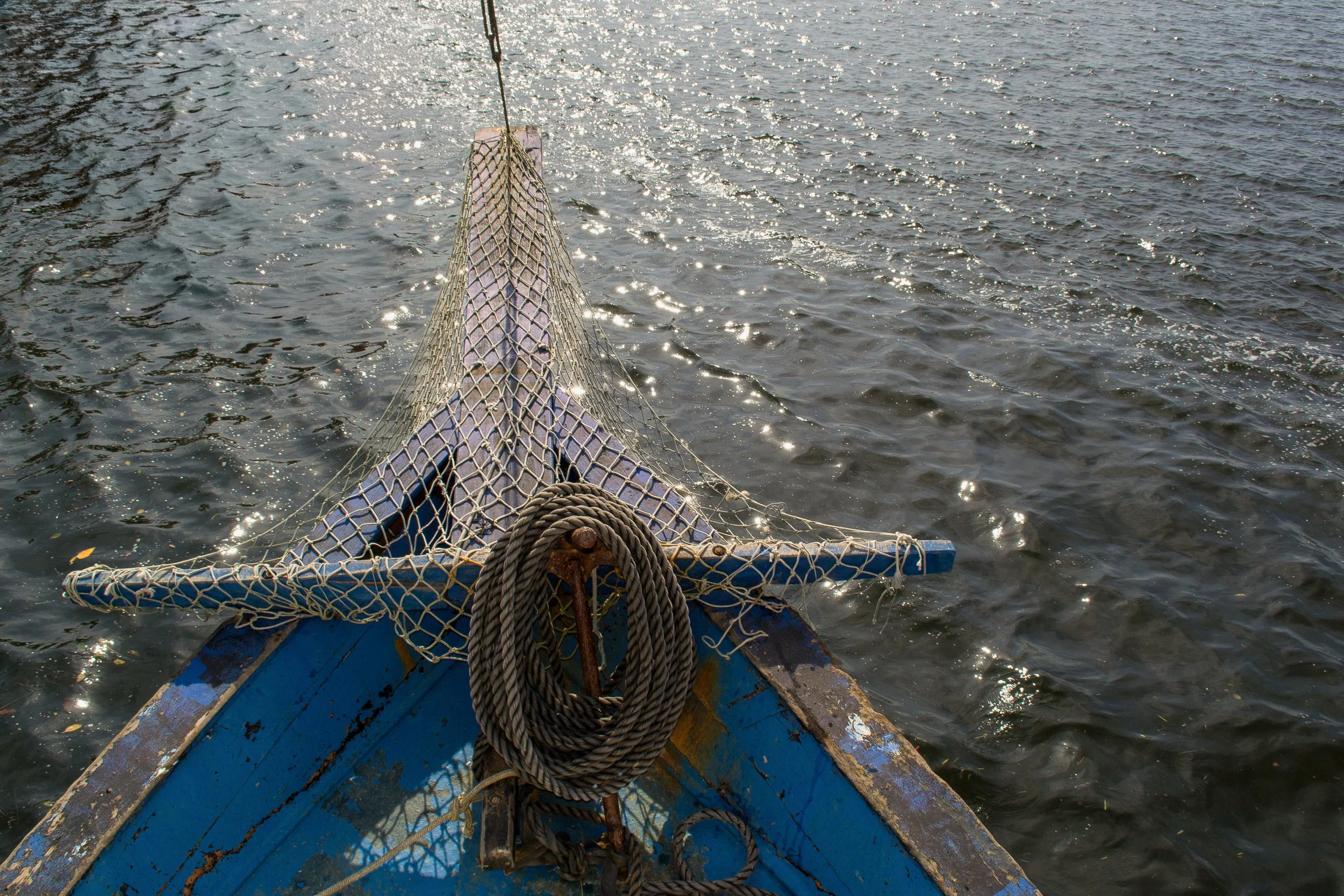 Proa de barco com redes de pesca, âncoras, e fita de amarração, navegando em águas calmas com reflexos do sol nas ondas.