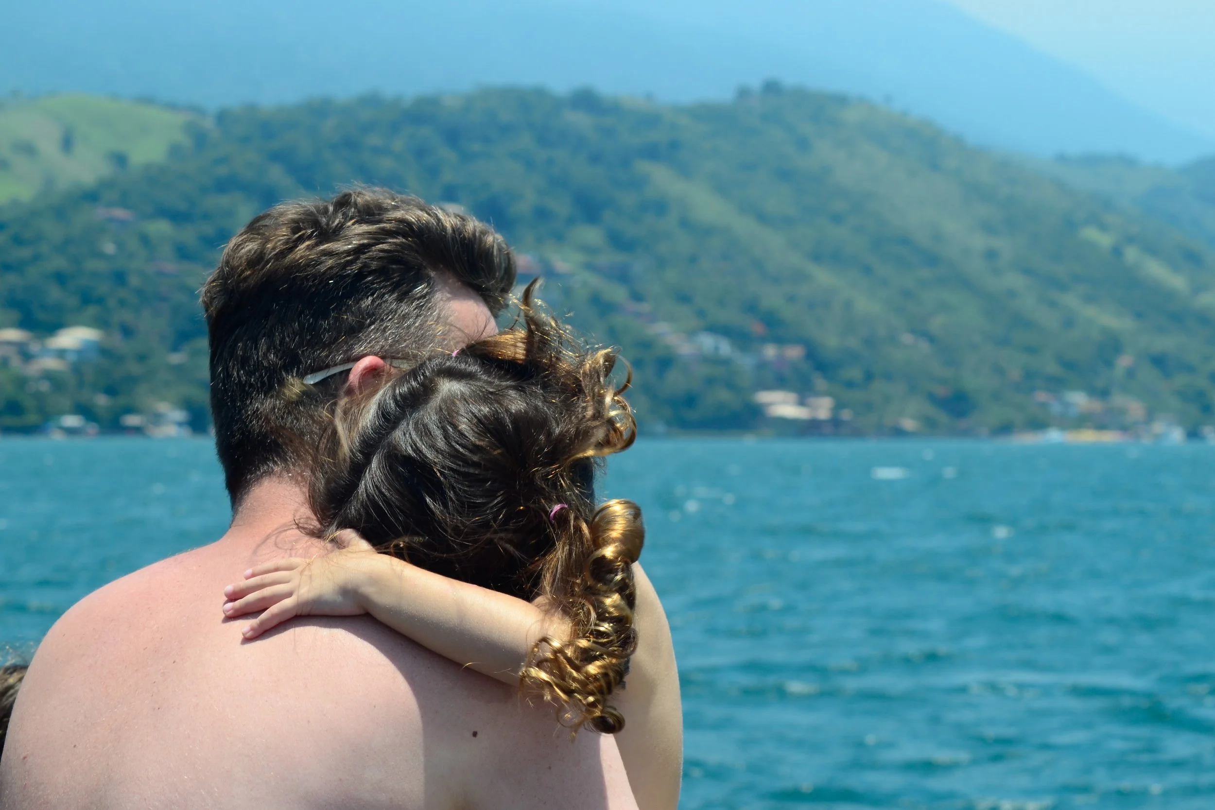 Casal abraçado em frente a um lago com montanhas no fundo.