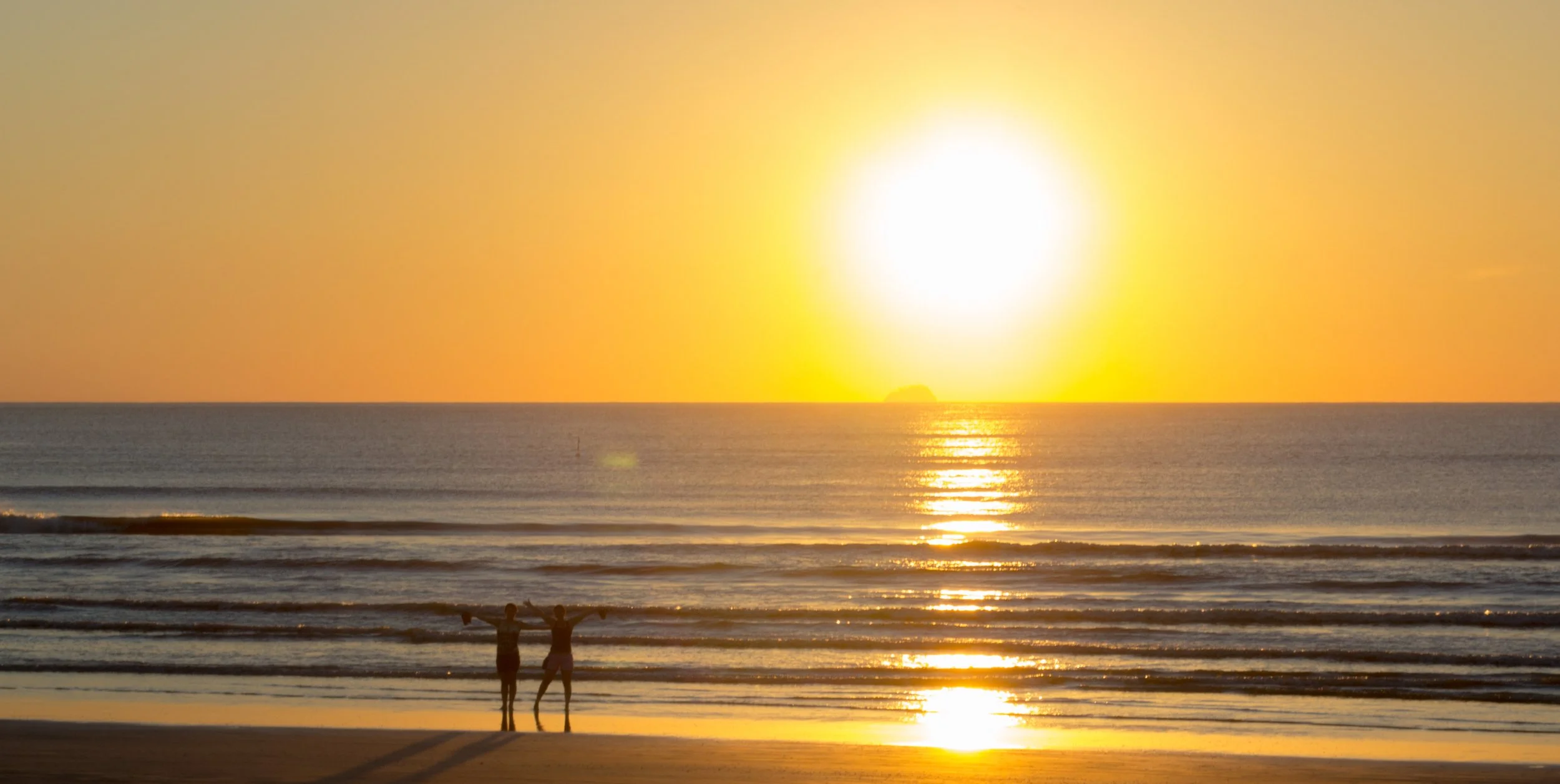 Pôr do sol na praia com duas pessoas com os braços abertos na beira da água.