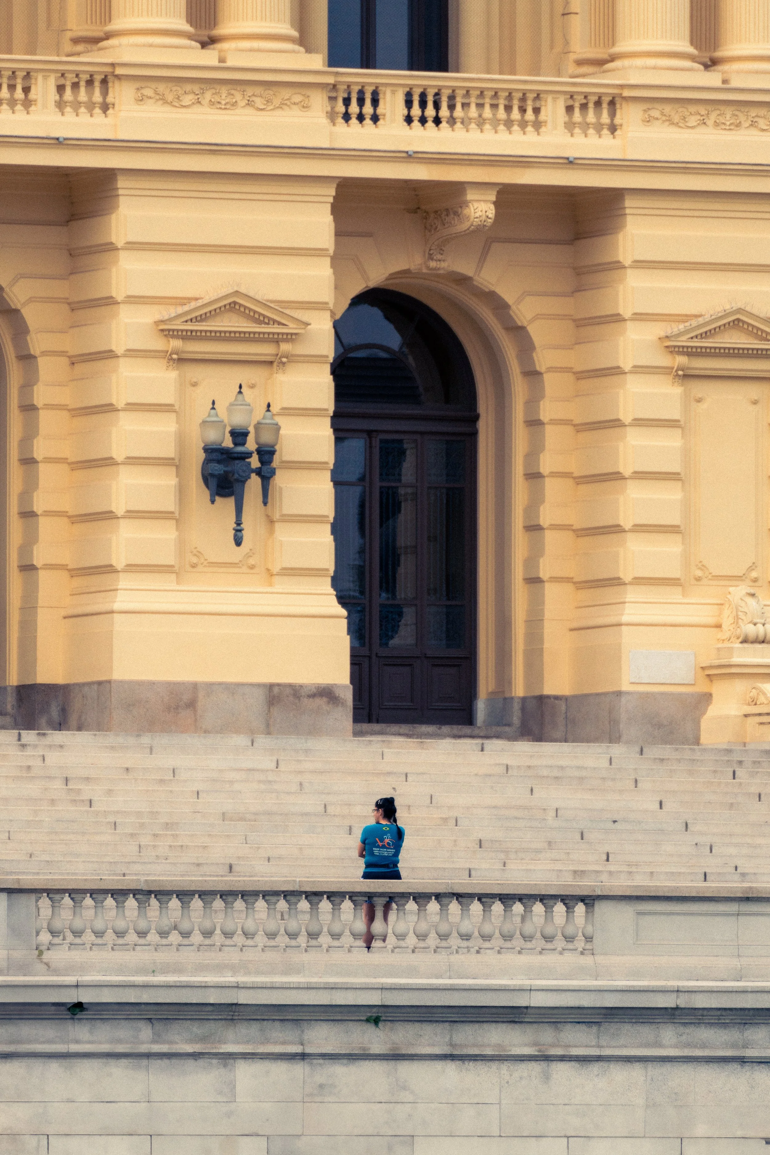 Pessoa de costas, usando blusa azul e shorts vermelhos, sentada nos degraus de um edifício clássico de cor amarela, com detalhes arquitetônicos ornamentados e iluminação de poste clássico ao lado.
