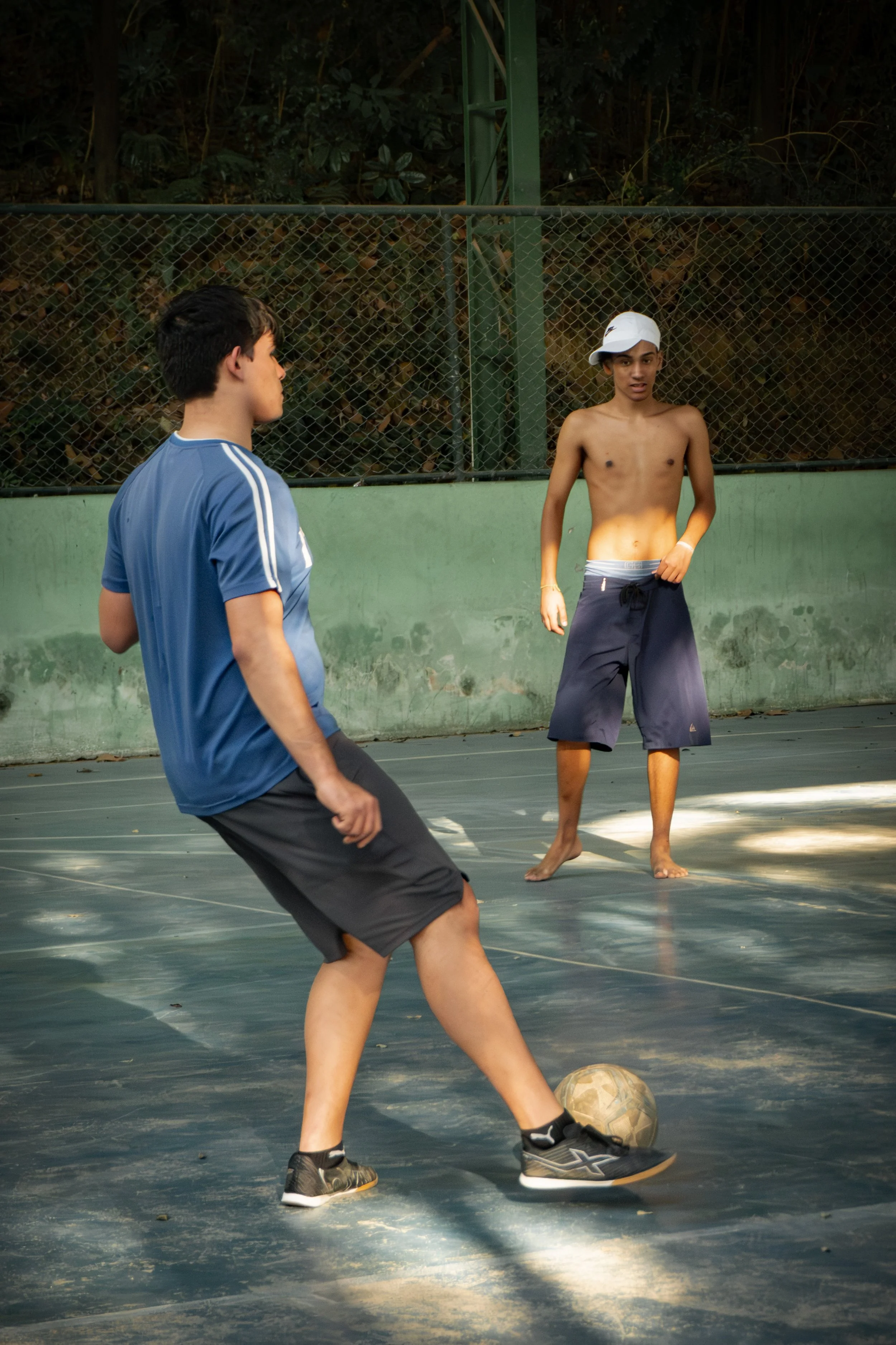 Jovens jogando futebol na quadra ao ar livre, um deles de camiseta azul e o outro sem camisa usando boné branco.