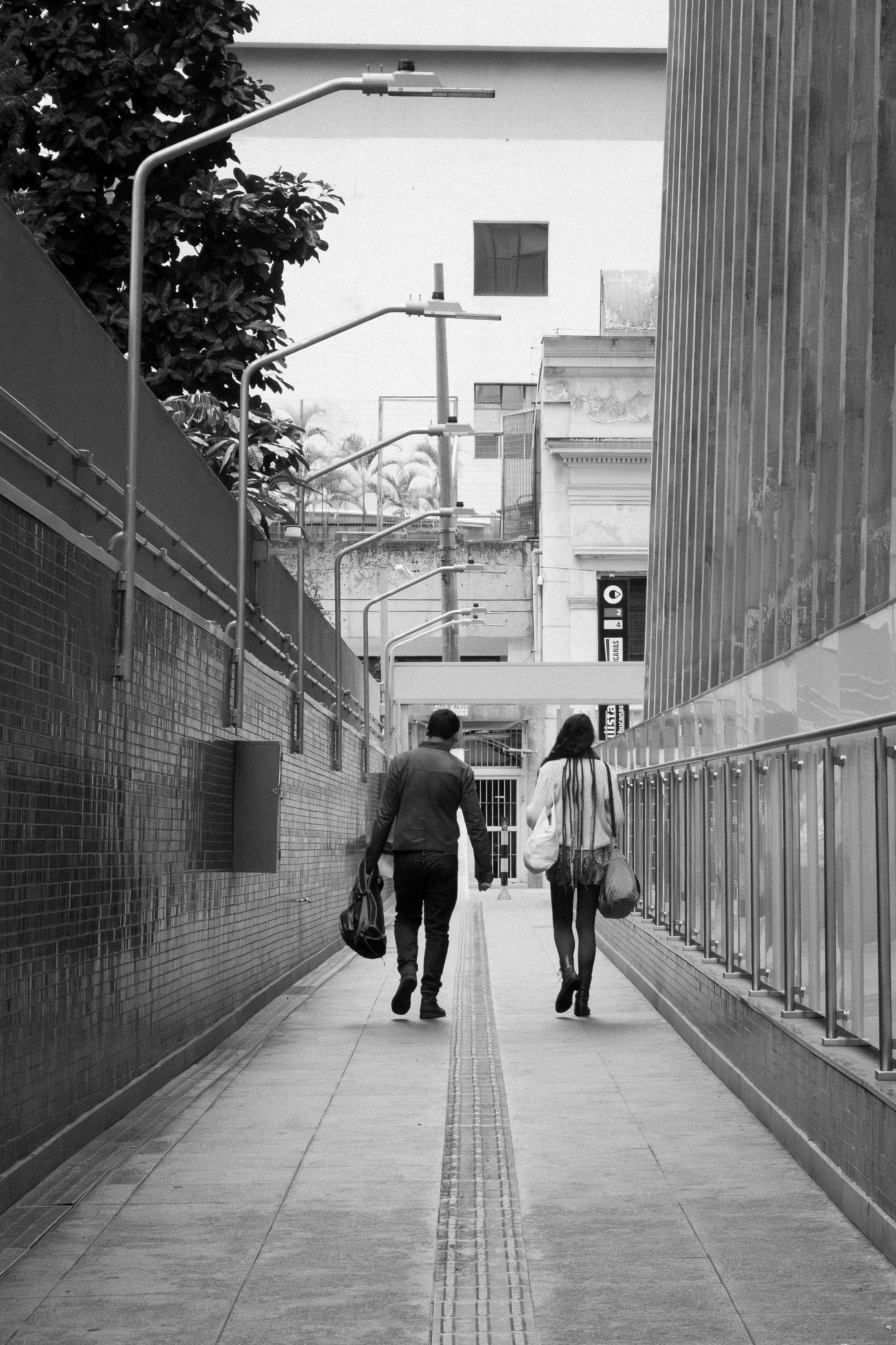 Imagem em preto e branco de um casal caminhando por uma passarela urbana entre dois prédios, com postes de iluminação ao lado esquerdo.