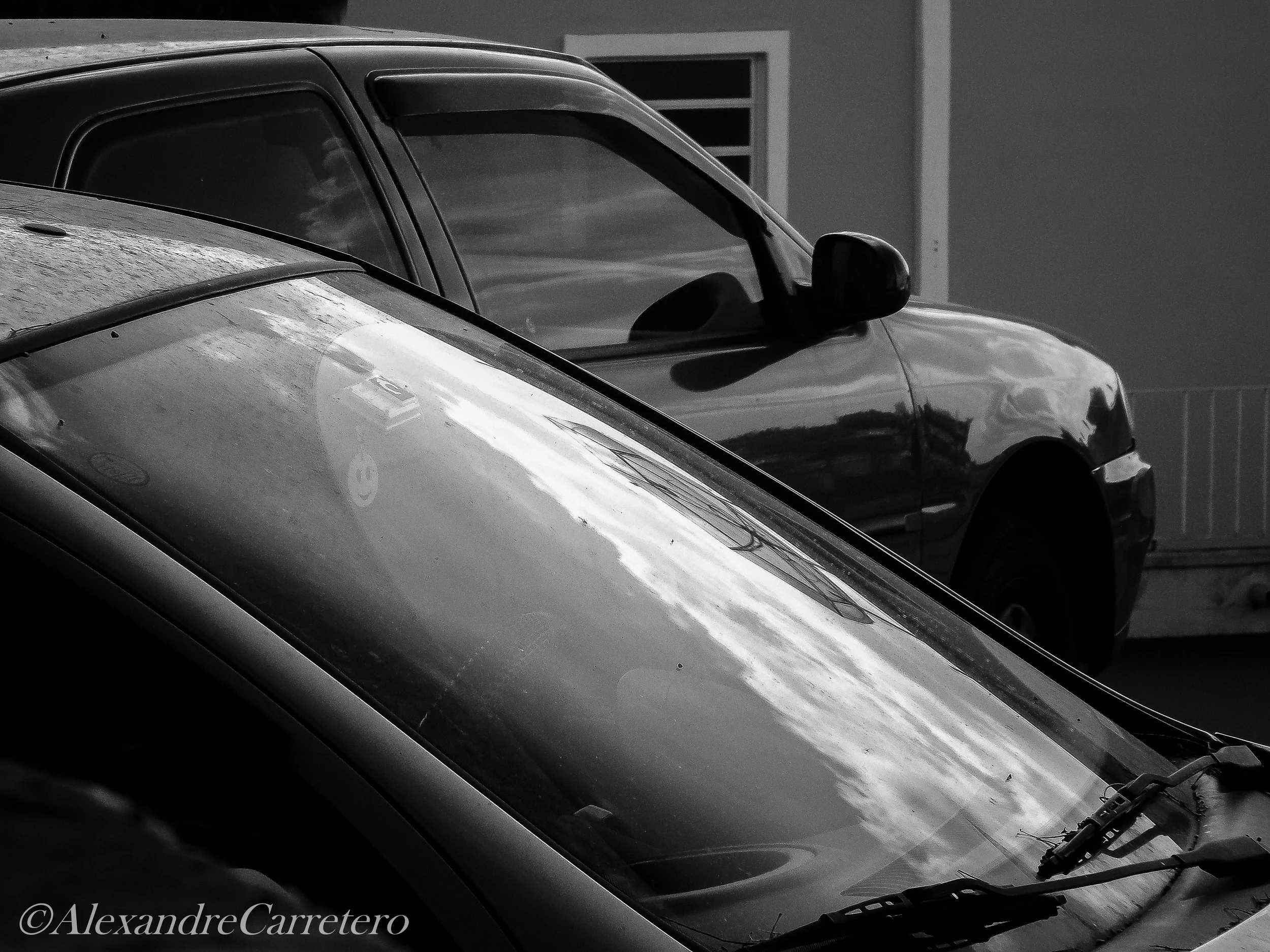 Dois carros estacionados, um em foco na frente, outro ao fundo, ambos em preto e branco, refletindo nuvens no vidro e nas superfícies.