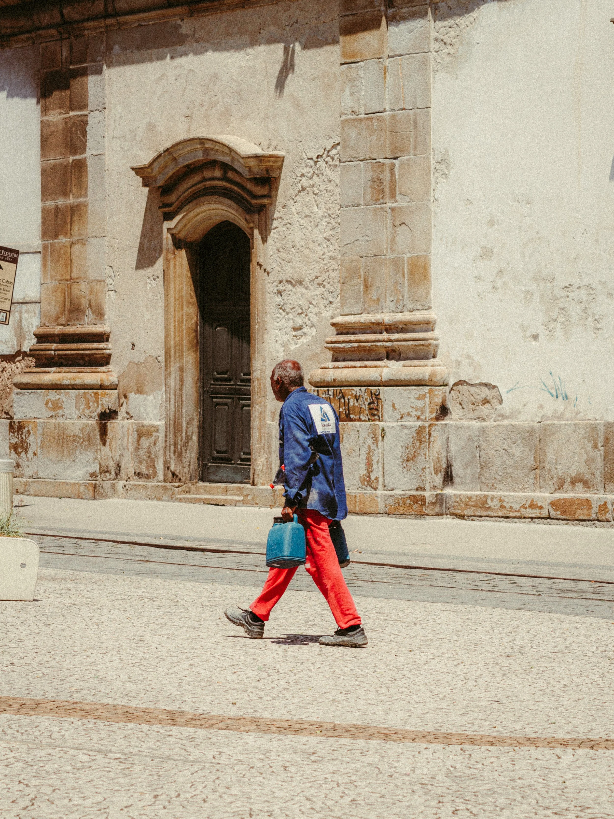 Homem atravessando a rua carregando uma mochila azul, com uma fachada antiga de pedra ao fundo.