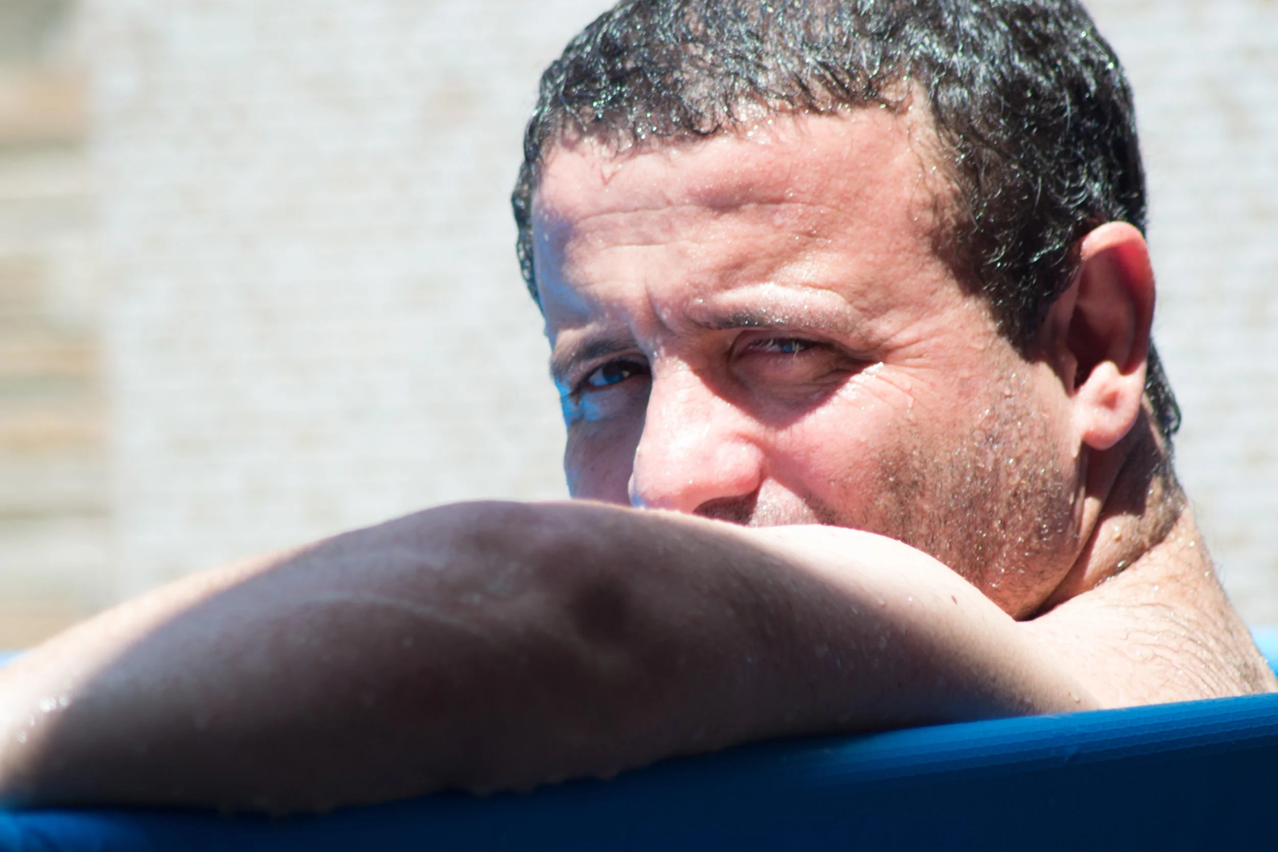 Homem com cabelo escuro, molhado, olhando de lado, descansando na borda de uma piscina com água clara ao fundo.