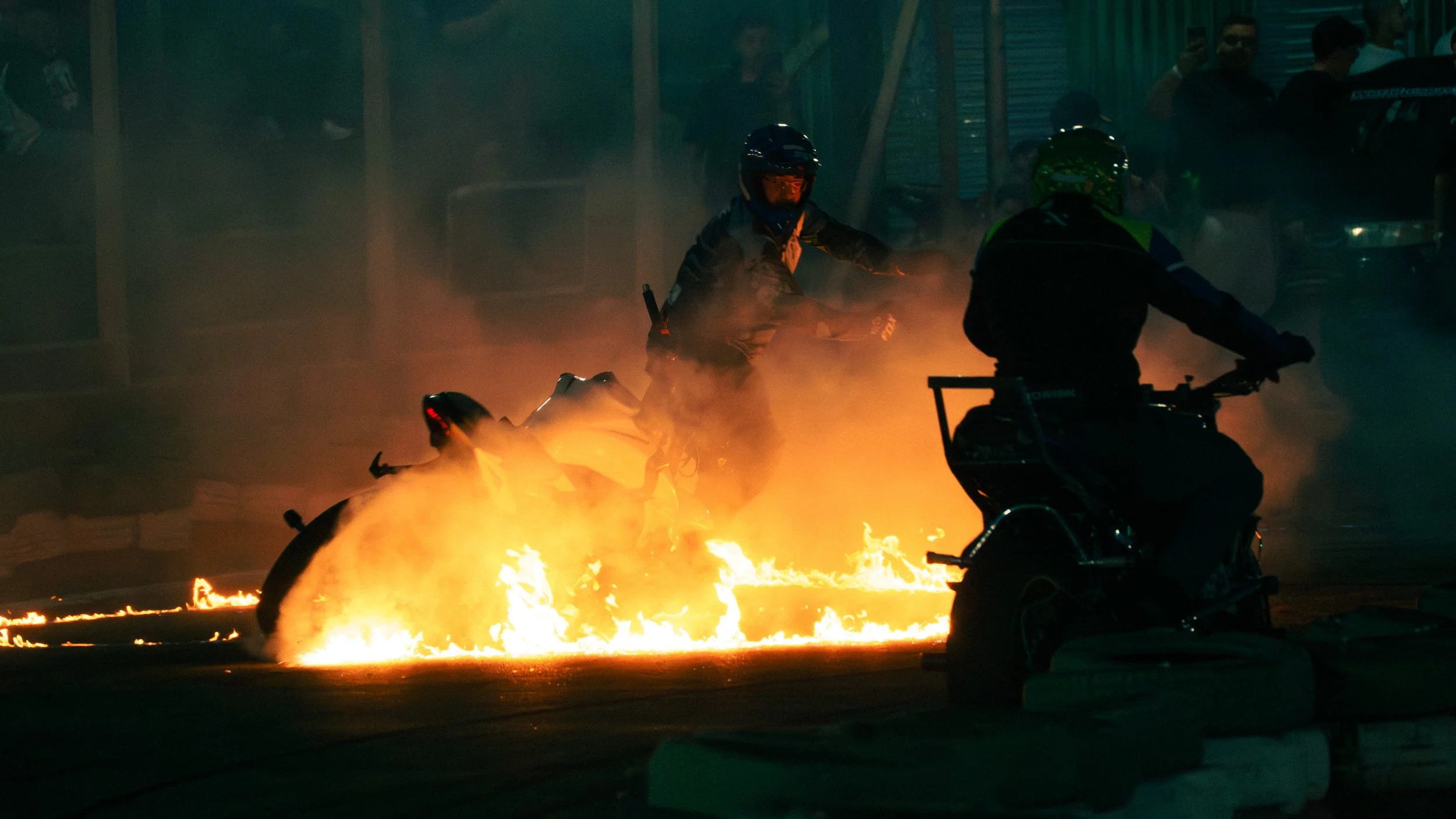 Duas pessoas em motos ao lado de uma fogueira na rua à noite, com fumaça ao redor.