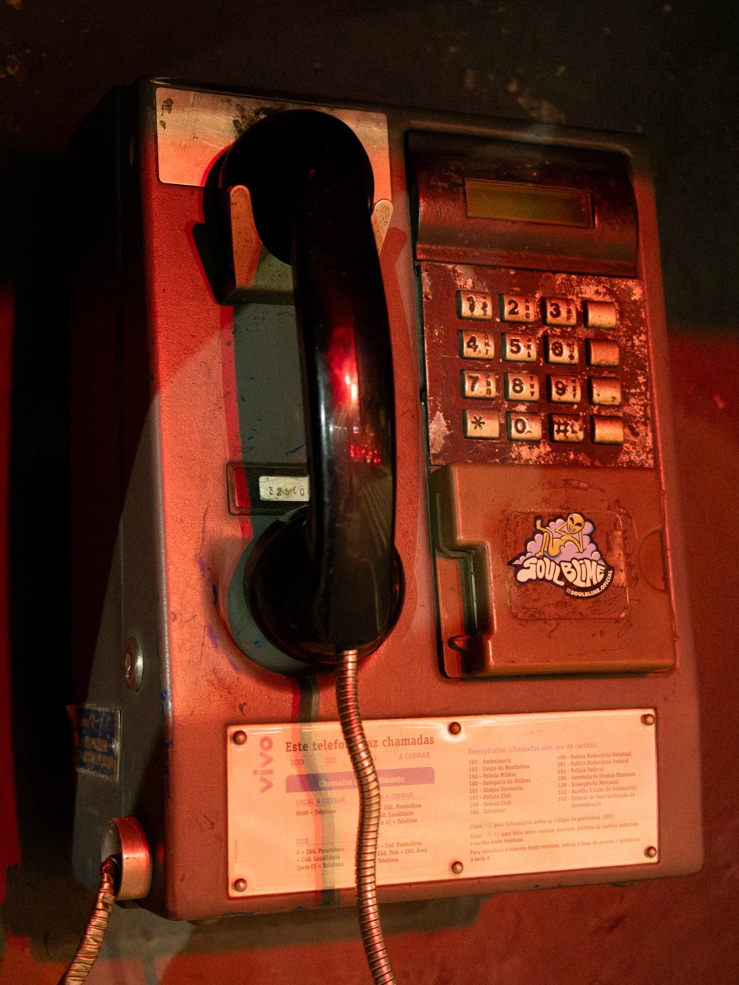 Telefone de praça antigo com teclado numérico, cabos enrolados, um adesivo cheio de desenhos e uma etiqueta de tarifas.