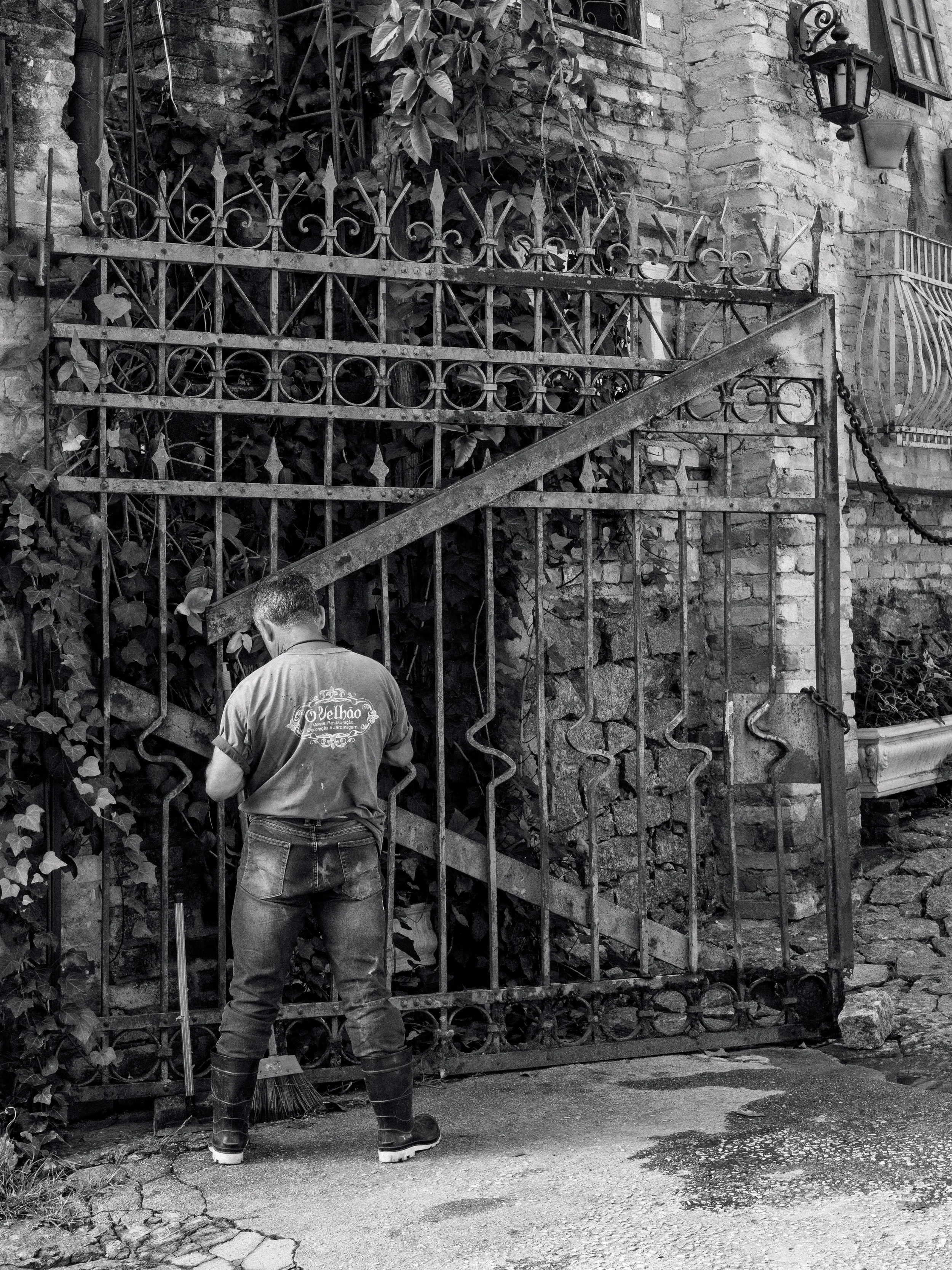 Pessoa limpando uma porta de ferro com uma escova, ao fundo uma parede de tijolos e plantas.