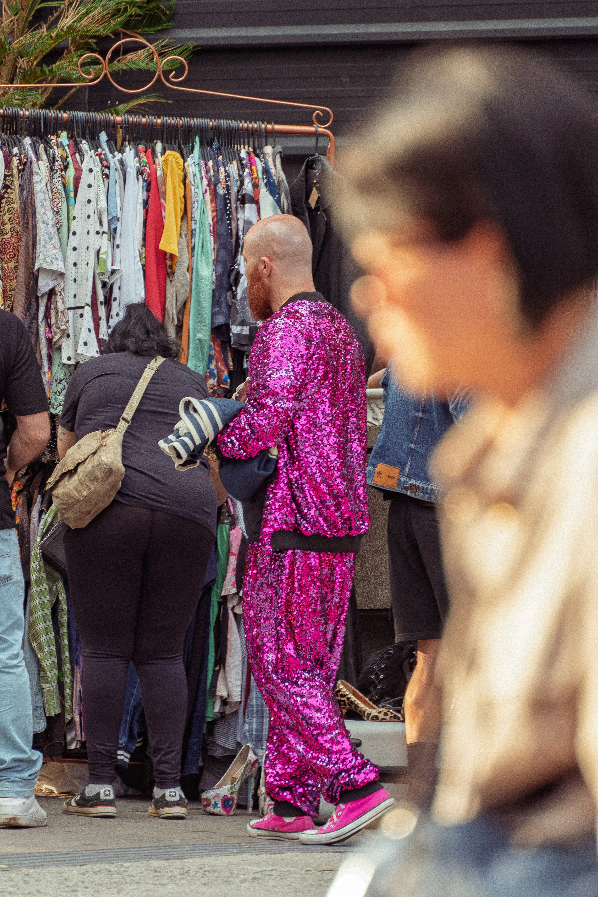 Homem com roupa brilhante rosa em uma feira de roupas com várias pessoas olhando roupas penduradas.